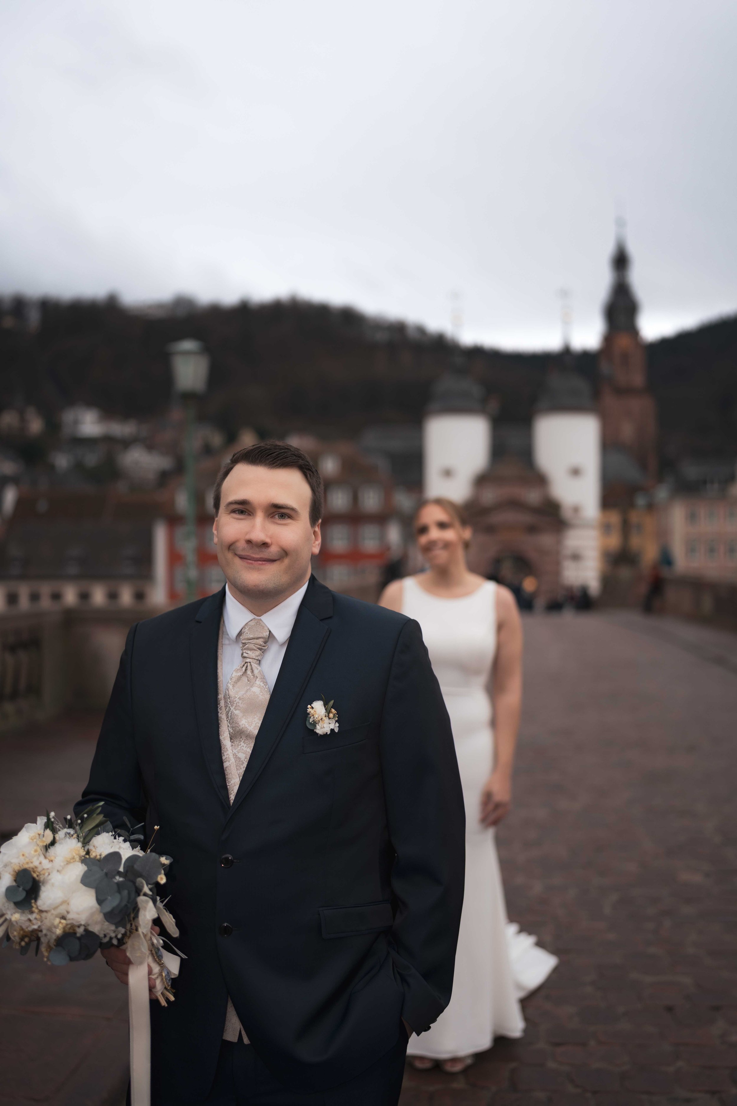 Ein Brautpaar steht auf einer Brücke in einer alten Stadt, mit einer Kirche im Hintergrund. Der Mann trägt einen schwarzen Anzug, und die Frau trägt ein weißes Brautkleid.