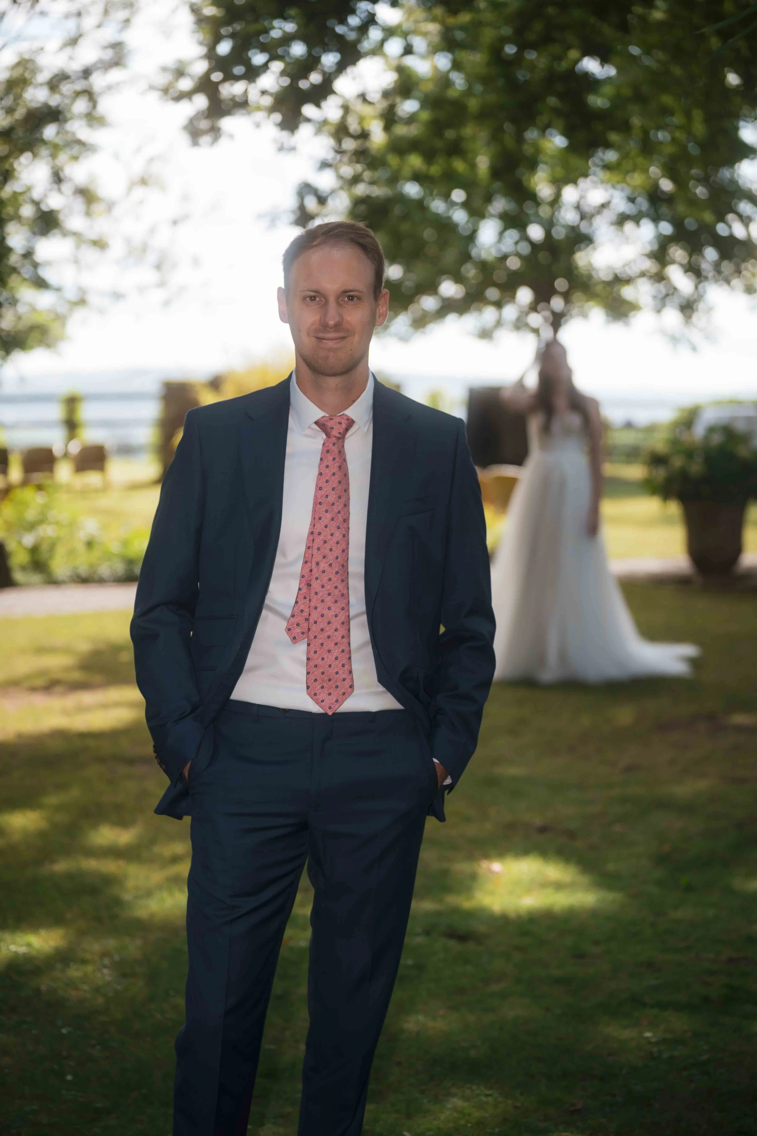 Ein Mann in Anzug mit Krawatte steht im Freien, im Hintergrund eine Frau in einem Hochzeitskleid unter einem Baum.