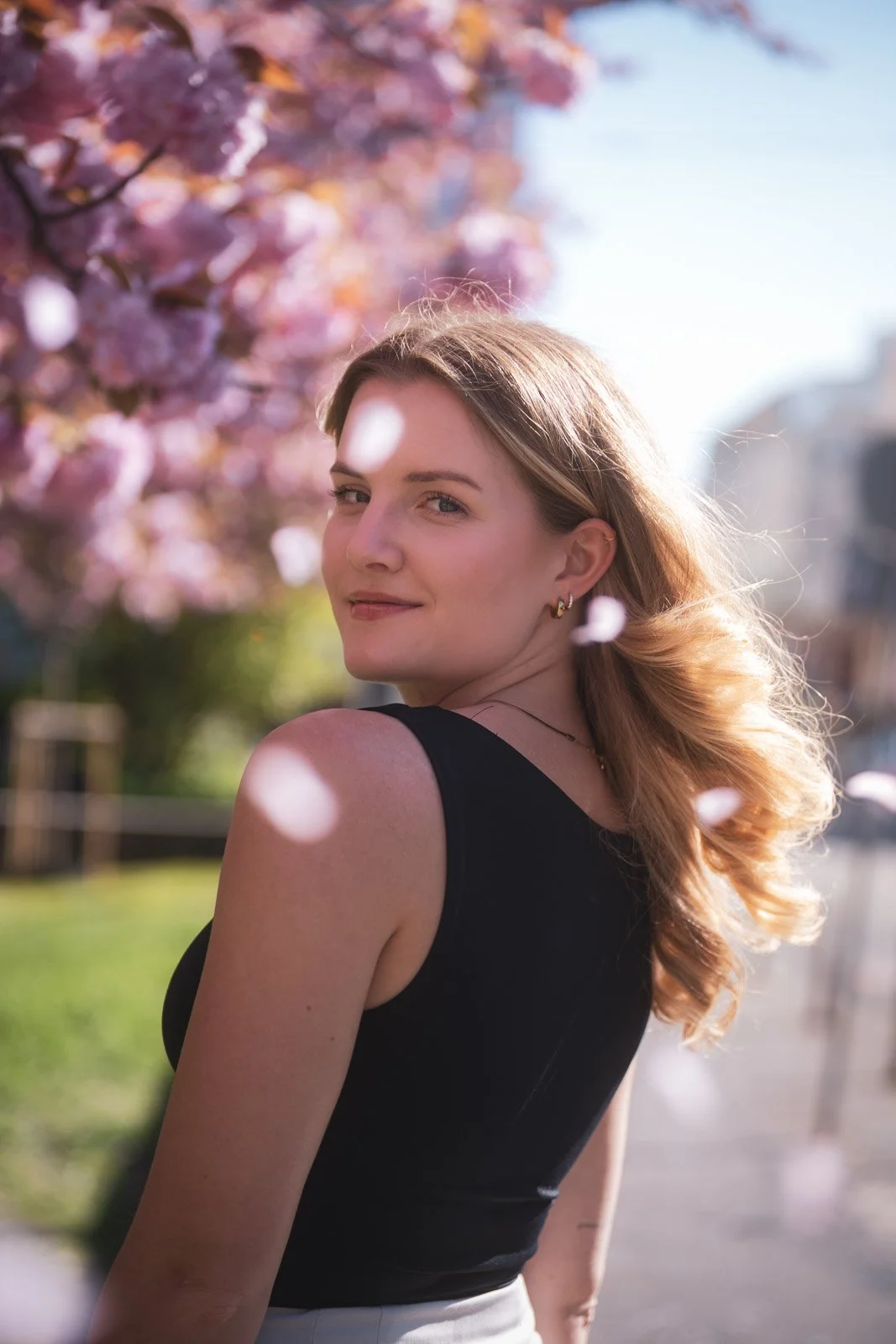 Junge Frau mit lockigem, blonden Haar, in Schwarz gekleidet, blickt lächelnd in die Kamera bei Sonnenlicht, vor rosa blühenden Bäumen im Freien.