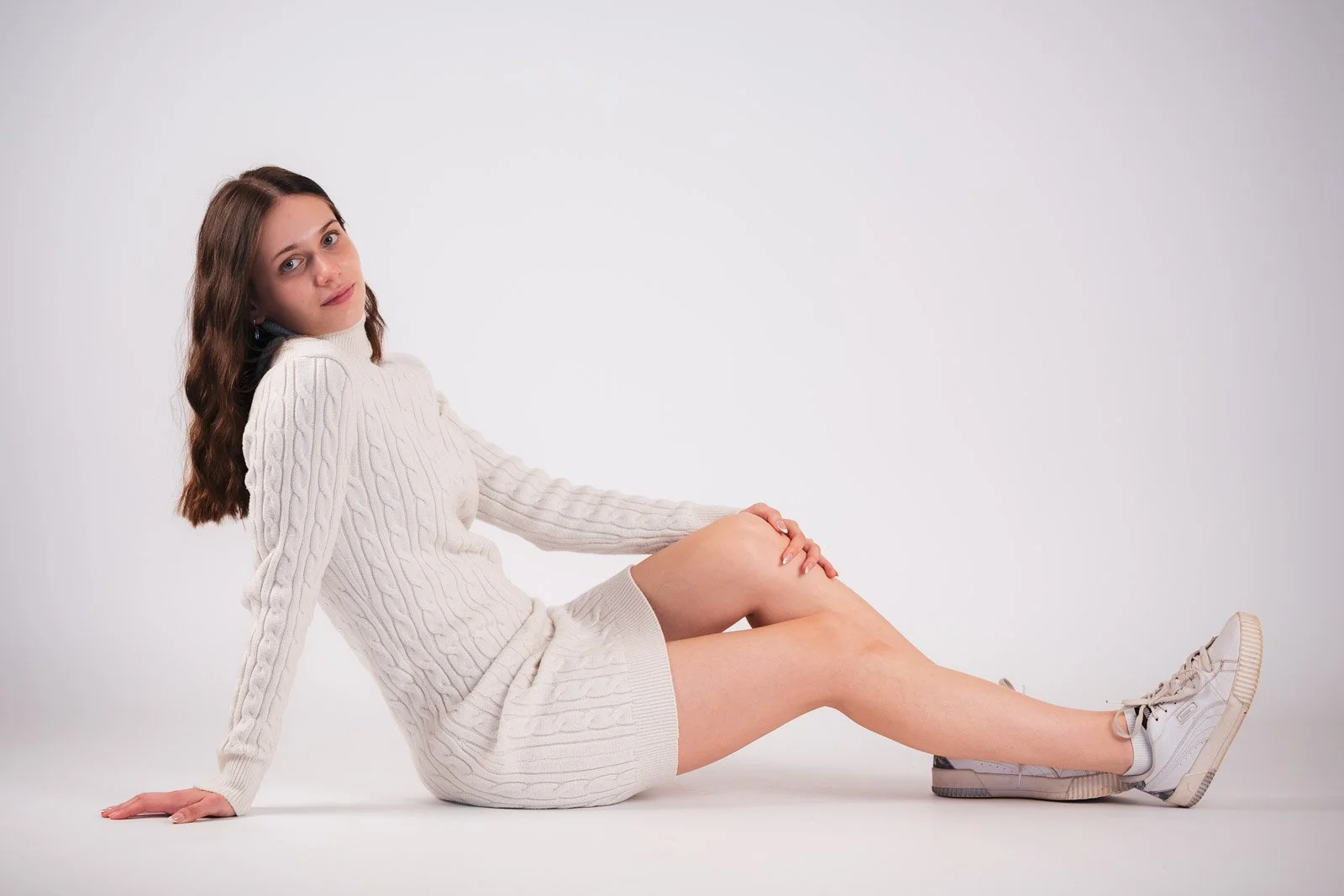 Junge Frau in einem weißen Strickpullover und Turnschuhen sitzt auf dem Boden, blickt in die Kamera, vor einem weißen Hintergrund.