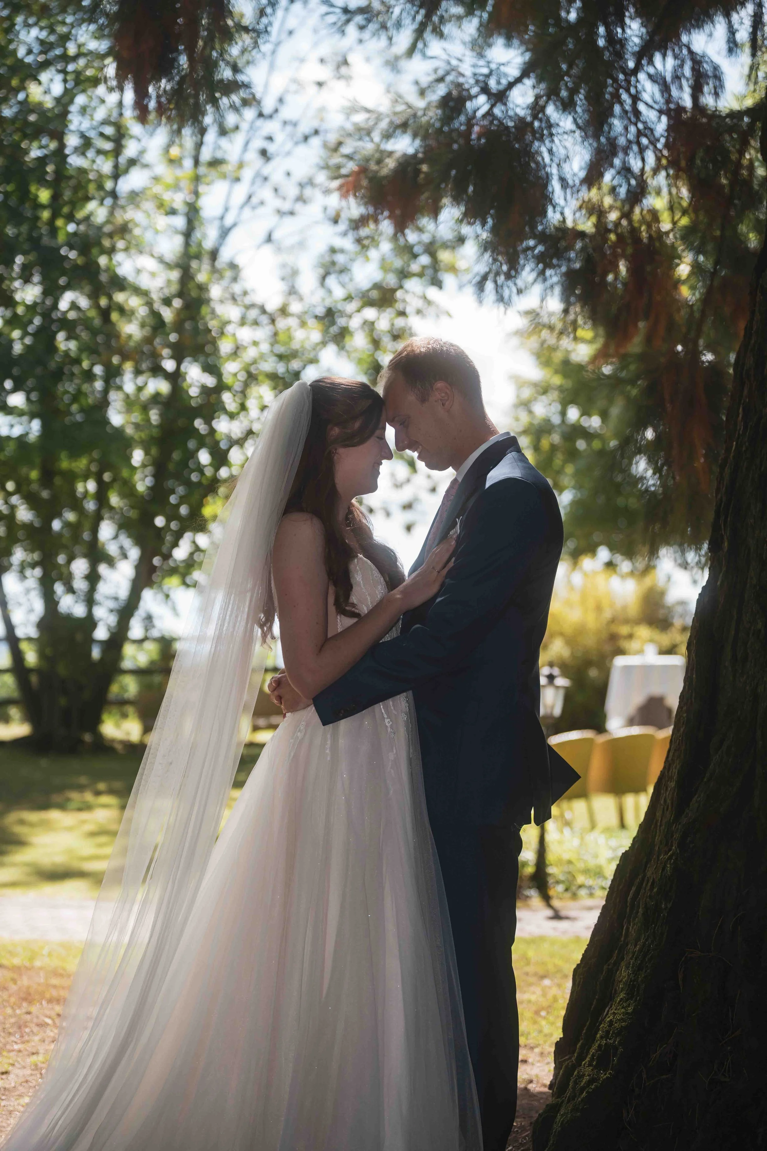 Braut und Bräutigam, die sich bei einer Hochzeit im Freien unter einem Baum gegenüberstehen, mit ihren Stirn- und Nasen zusammen. Sie sind in formeller Kleidung, mit dem Brautkleid inklusive Schleier und dem Anzug des Bräutigams.