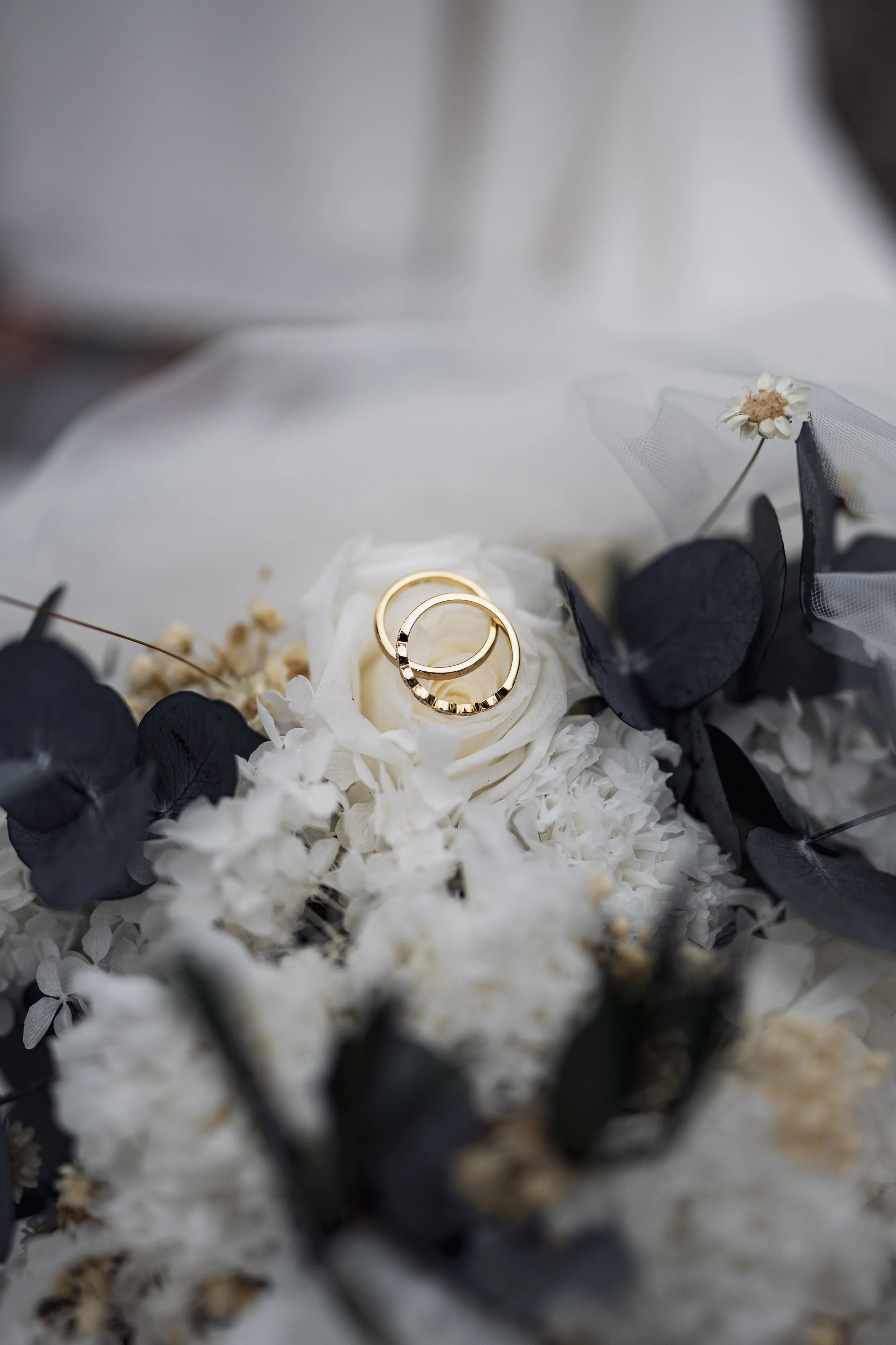 Zwei goldene Eheringe liegen auf weißen Blumen in einem Brautstrauß.