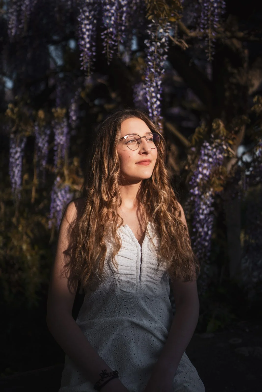 Junge Frau mit langen, lockigen Haaren und Brille trägt ein weißes Kleid und steht vor einer blühenden Fliederhecke bei Sonnenuntergang.