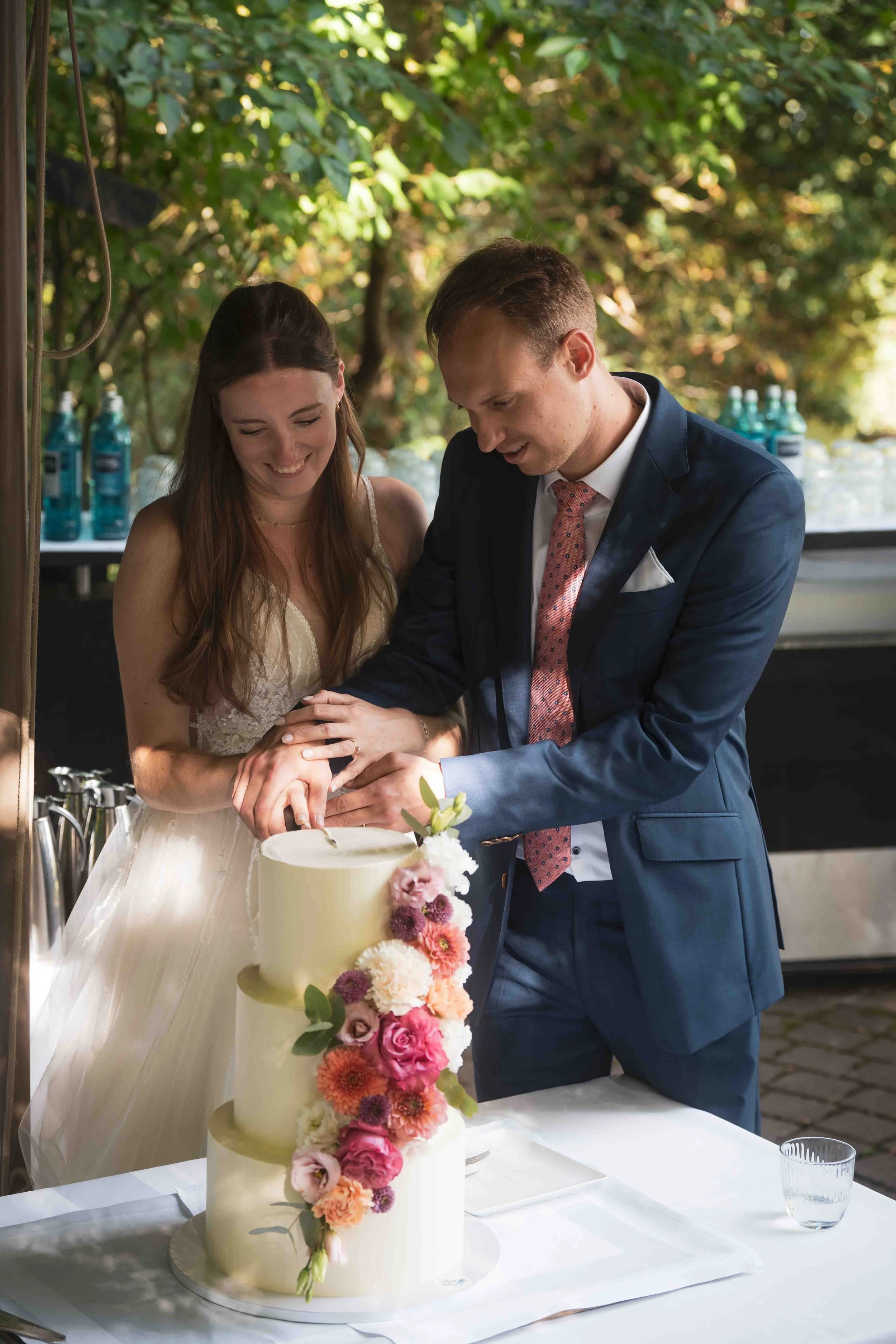 Ein Paar schneidet eine Hochzeitstorte bei einer Feier im Freien, mit einer zweistöckigen Torte mit Blumenverzierung.