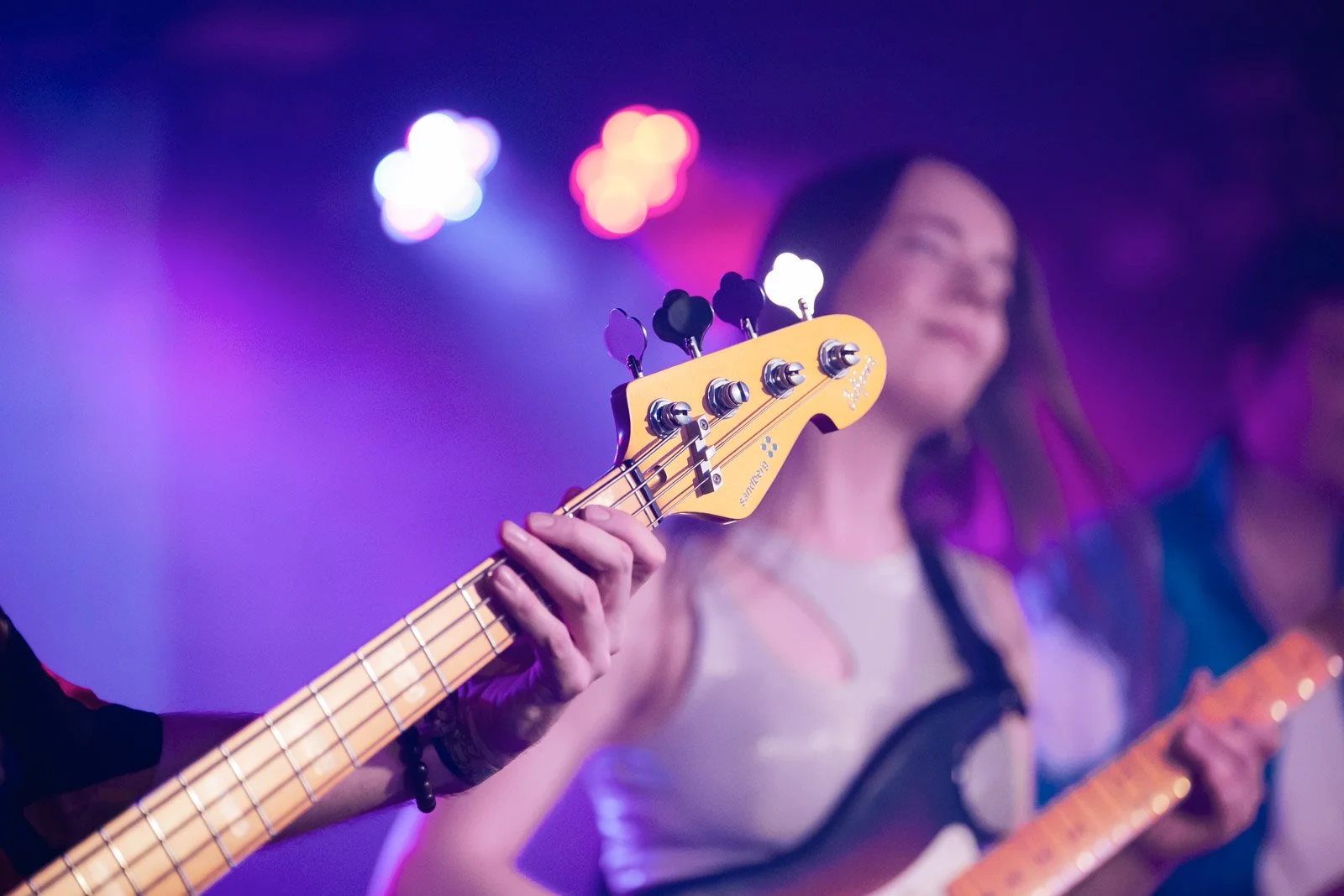 Nahaufnahme eines Gitarrenkopfes mit vier Tasten und einer Person im Hintergrund beim Gitarrespielen bei einem Musikauftritt, mit bunter Bühnenbeleuchtung.