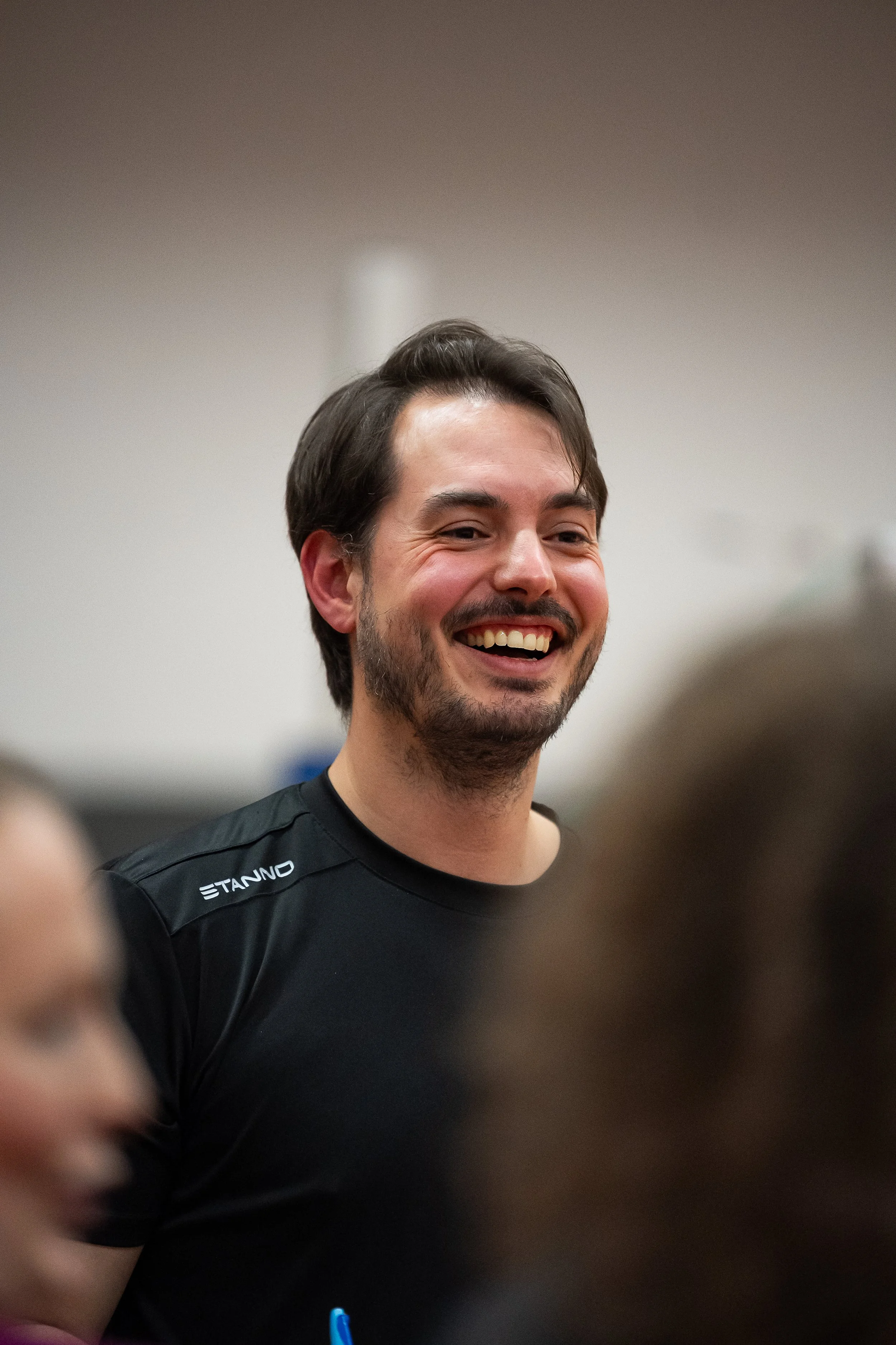 Ein lachender junger Mann mit dunklem Haar und Bart, trägt ein schwarzes T-Shirt mit dem weißen Logo 'ETANNO', steht in einem Raum mit weißer Wand im Hintergrund.