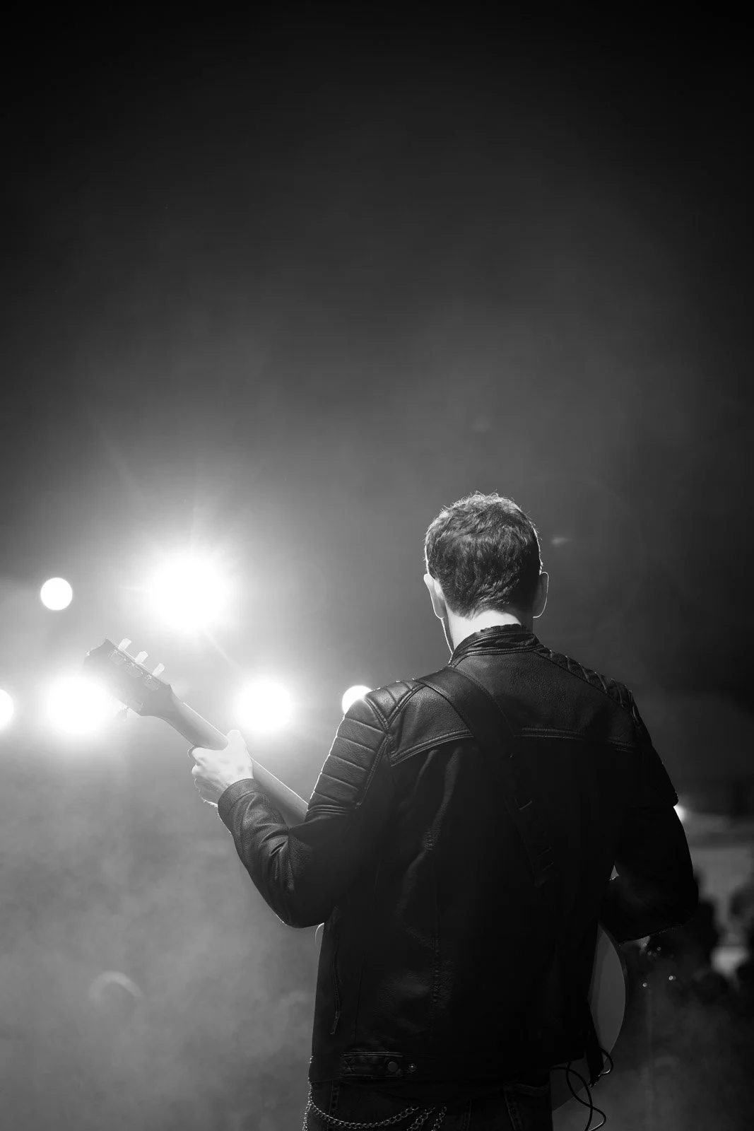 Schwarz-weiß-Foto eines Mannes mit einem E-Gitarre, der auf einer Bühne bei einem Konzert spielt, mit Scheinwerfern im Hintergrund.