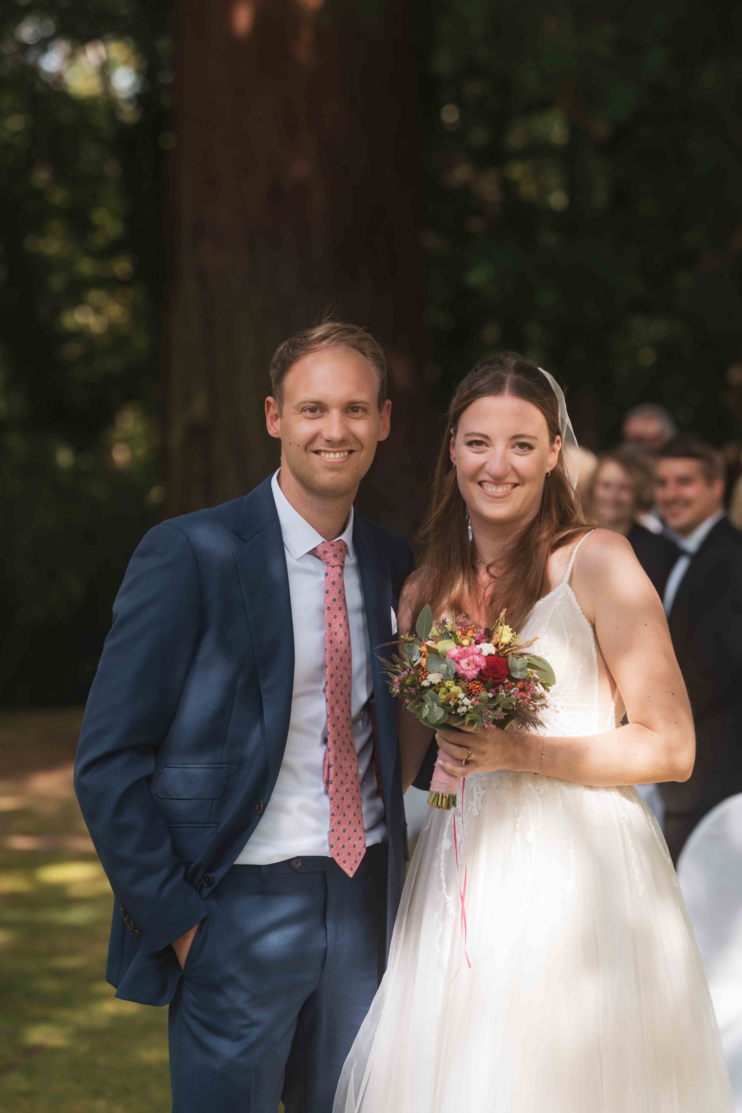 Ein Brautpaar bei einer Hochzeit im Freien, die Frau trägt ein weißes Brautkleid und hält einen Blumenstrauß, der Mann trägt einen dunkelblauen Anzug mit rosa Krawatte.