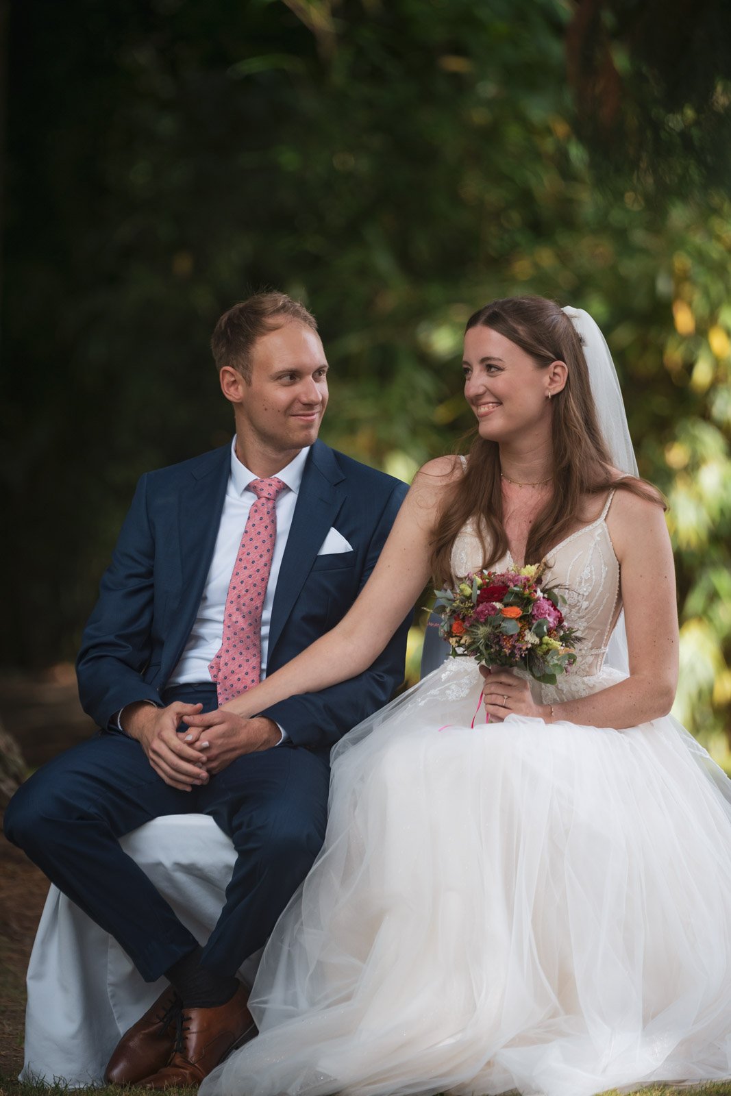 Ein Brautpaar sitzt auf einem Stuhl in einem Wald, der Bräutigam trägt einen dunkelblauen Anzug mit roter Krawatte, die Braut trägt ein weißes Brautkleid und hält einen Blumenstrauß.