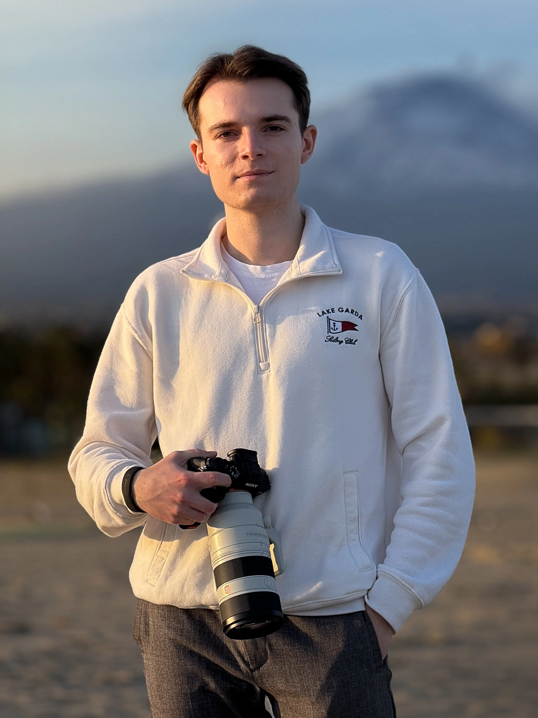 Junge Mann steht im Freien, trägt eine weiße Jacke mit 'Lake Garda' Stickerei, hält eine Kamera mit großem Objektiv, im Hintergrund Berge und ein bewölkter Himmel.