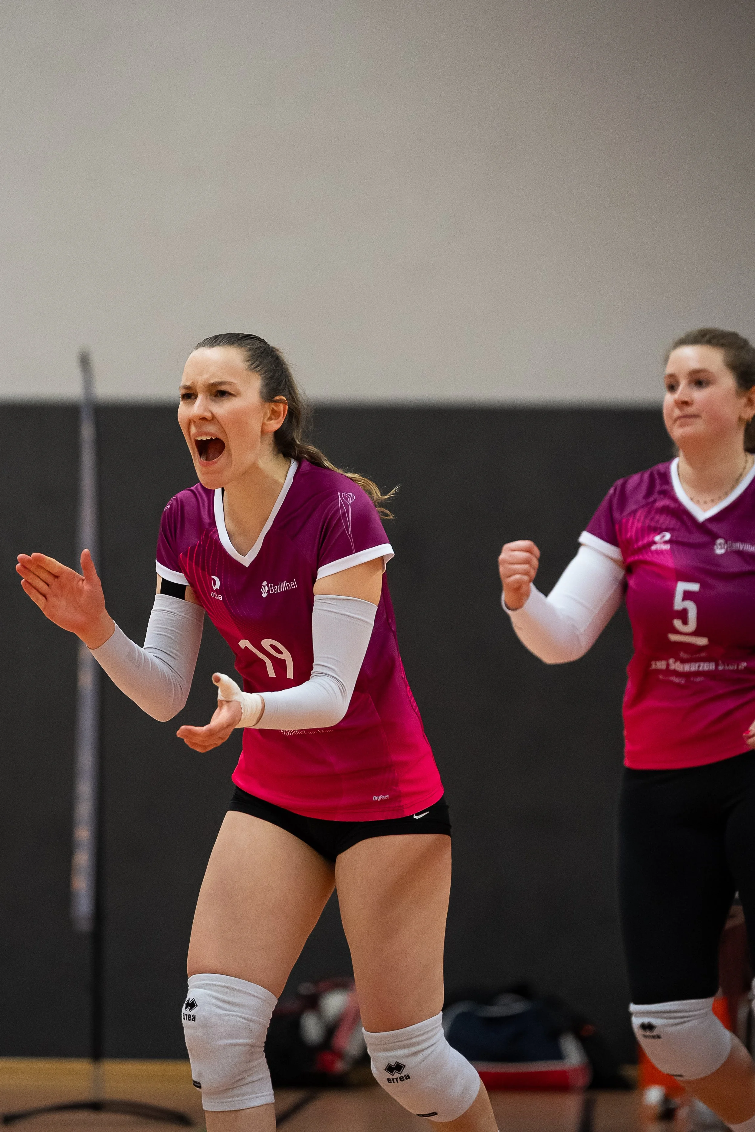 Zwei Frauen spielen Volleyball, tragen pinkfarbene Trikots mit weißen Ärmeln und schwarzen Shorts, eine jubelt oder schreit, während die andere auf der rechten Seite in einer kämpferischen Haltung steht.