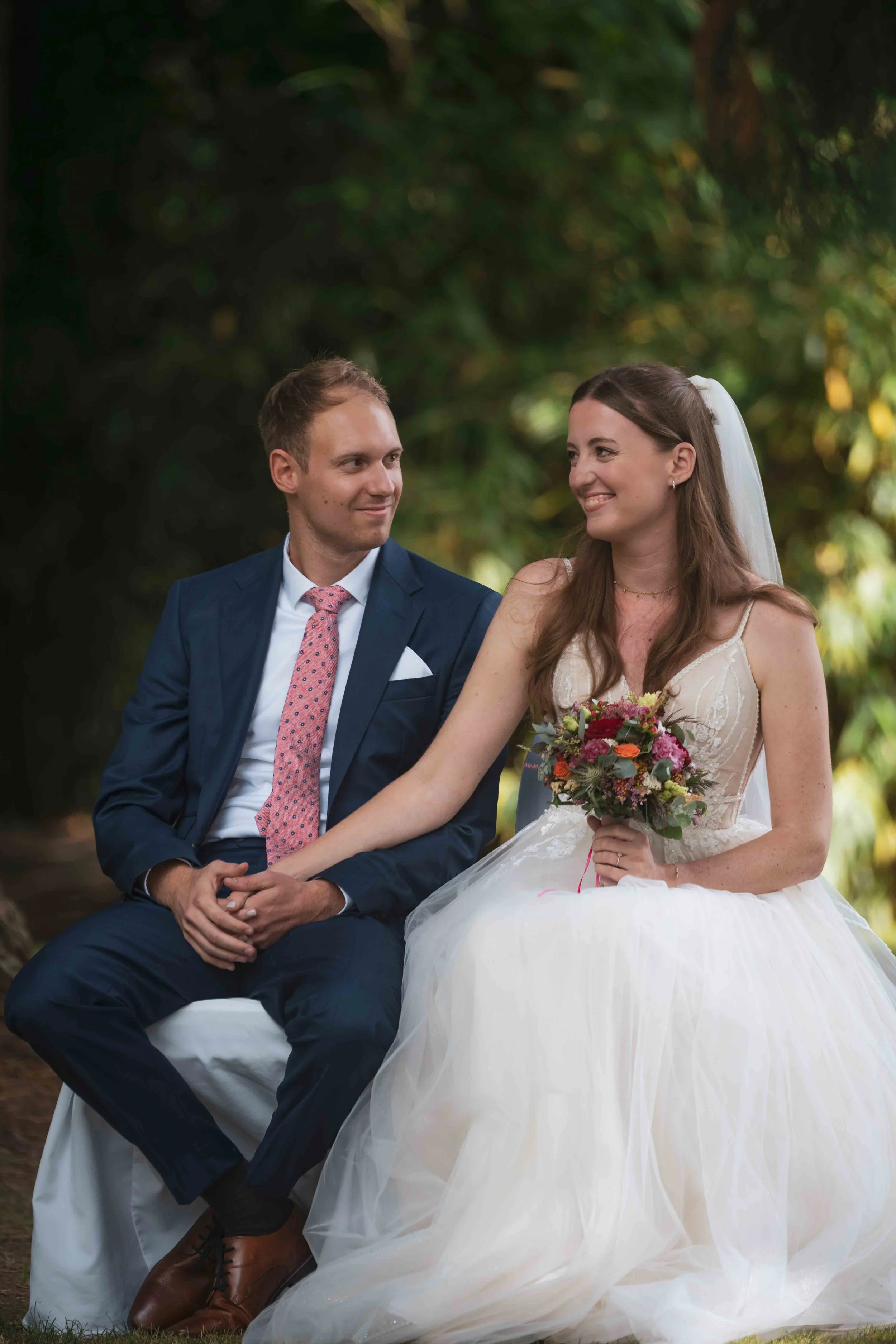 Ein Brautpaar sitzt gemeinsam unter einem Baum, die Braut trägt ein weißes Kleid und hält einen Blumenstrauß, der Bräutigam trägt einen dunkelblauen Anzug mit roter Krawatte.