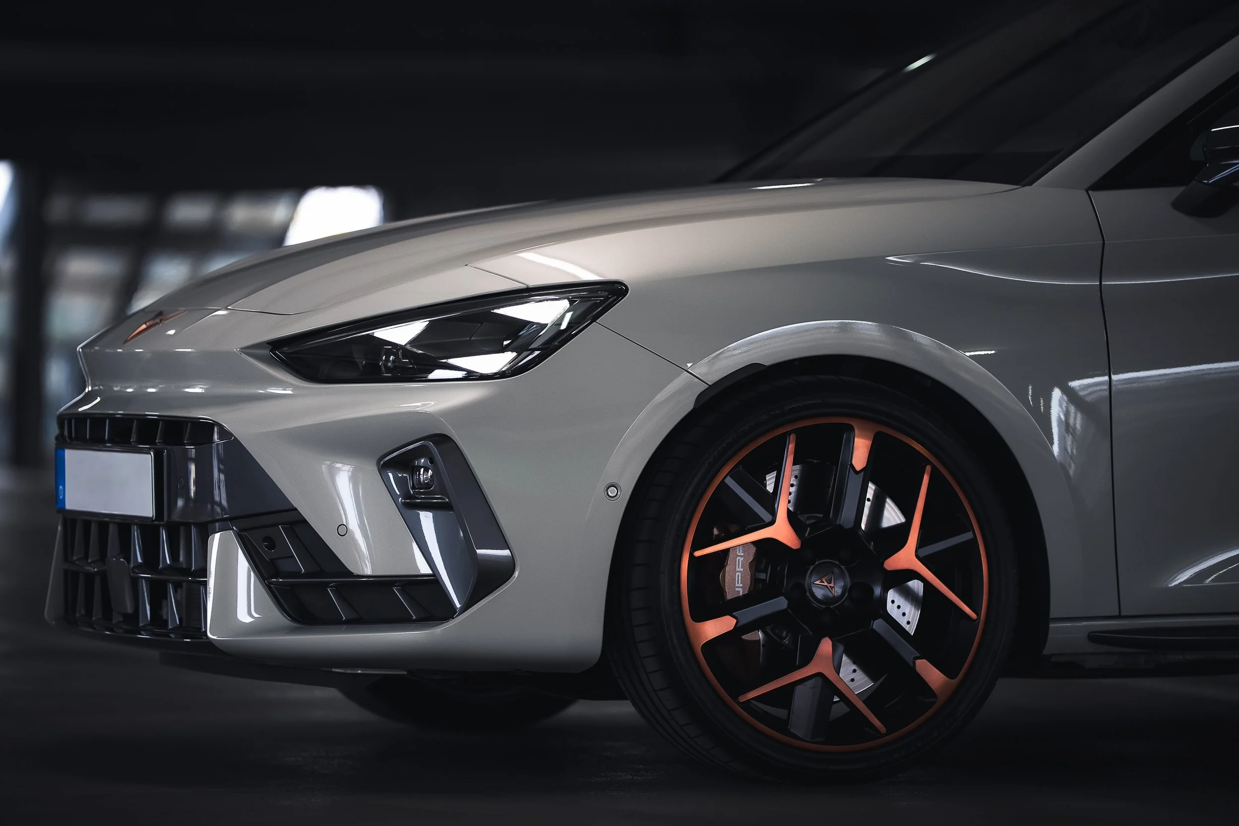 Vordere Fahrzeugfront eines modernen grauen Sportwagens mit schwarzen und orangefarbenen Felgen in dunkler Garage.
