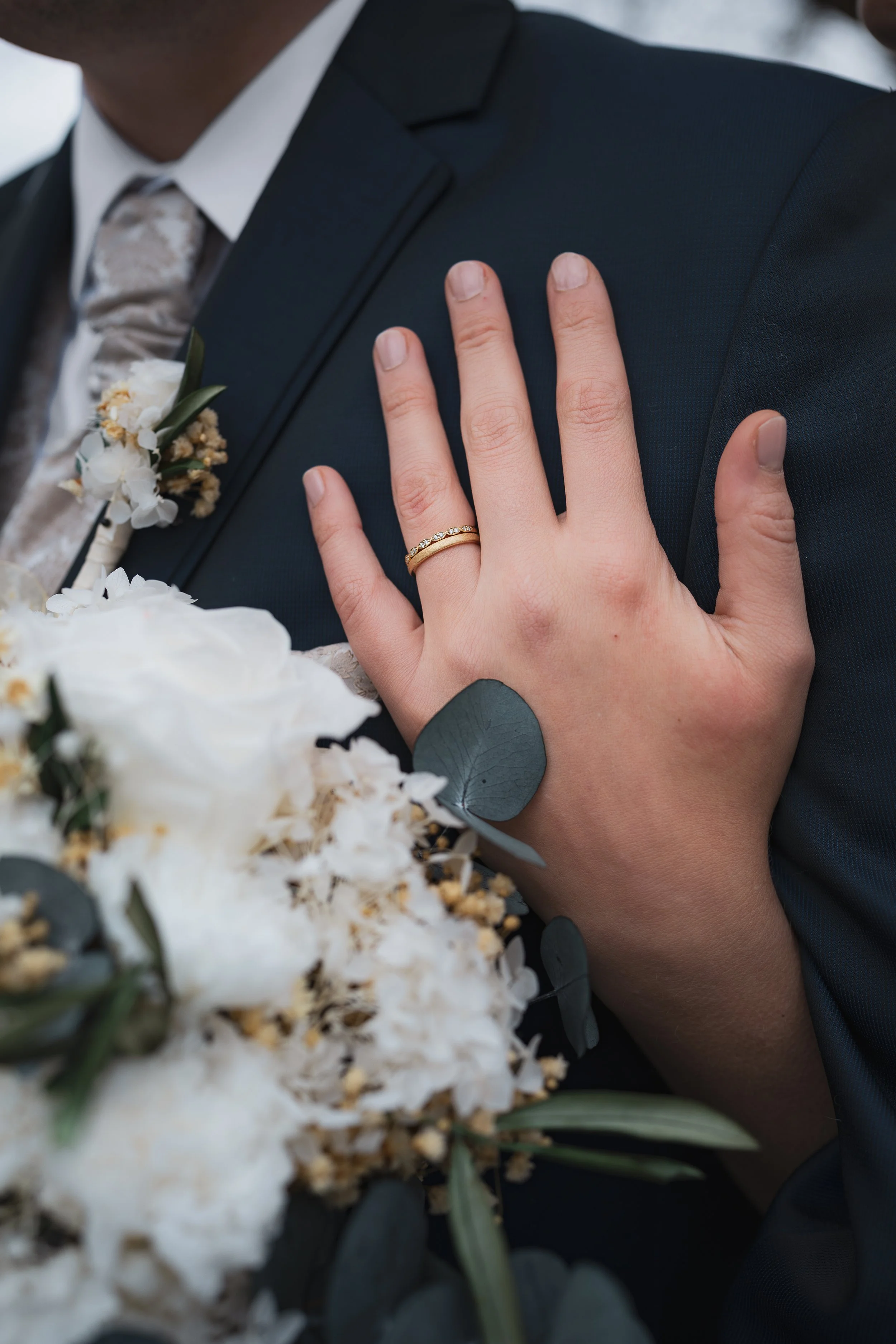 Nahaufnahme eines Paares bei einer Hochzeit, mit Fokus auf die Hand der Braut, die eine goldene Ehering trägt, auf die Schulter des Bräutigams gelegt, umgeben von weißen Blumen und Eukalyptusblättern.
