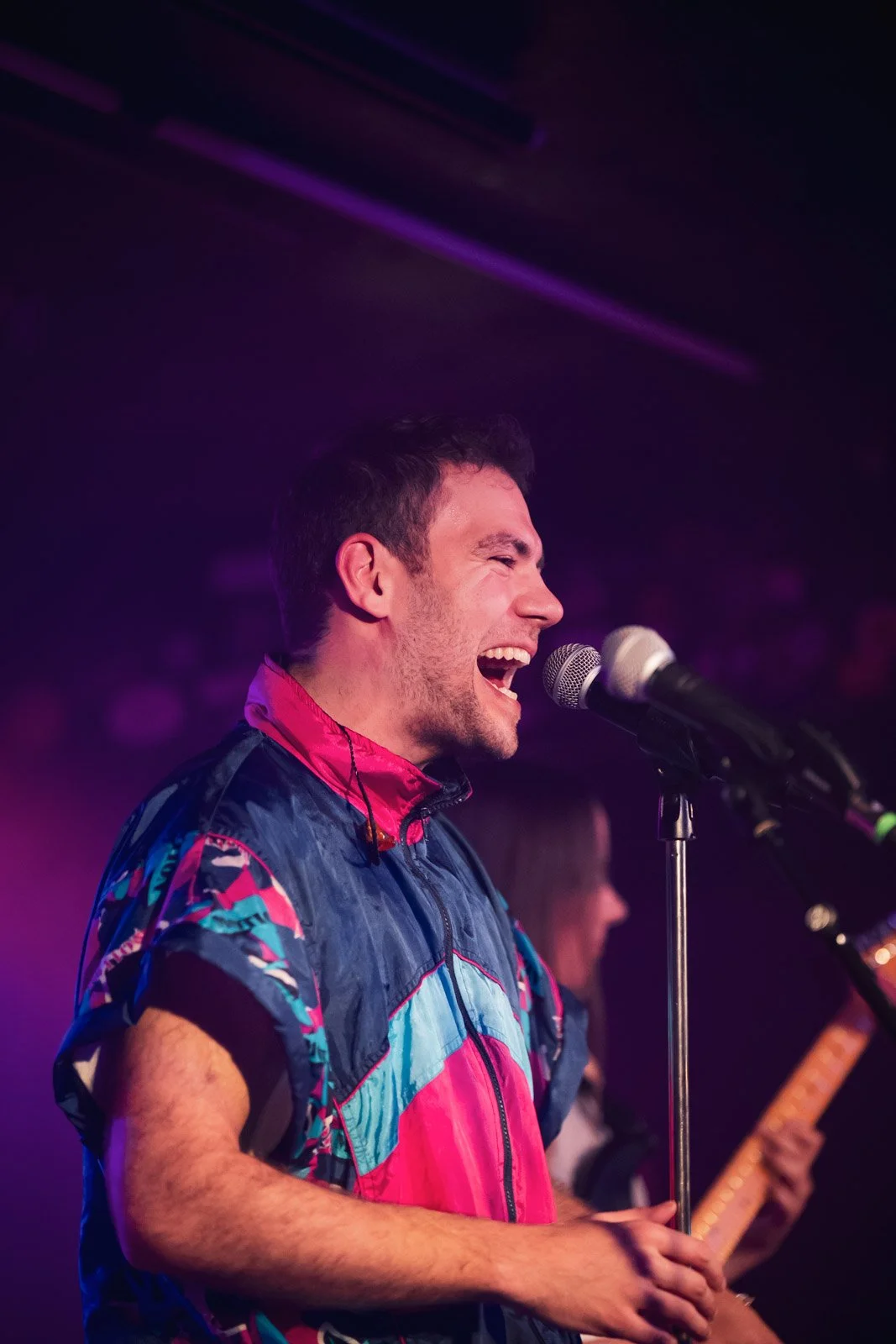 Ein junger Mann singt leidenschaftlich in ein Mikrofon auf einer Bühne, im Hintergrund ist eine Gitarre zu sehen, bei einer Live-Mus performance, mit farbigem Bühnenlicht.