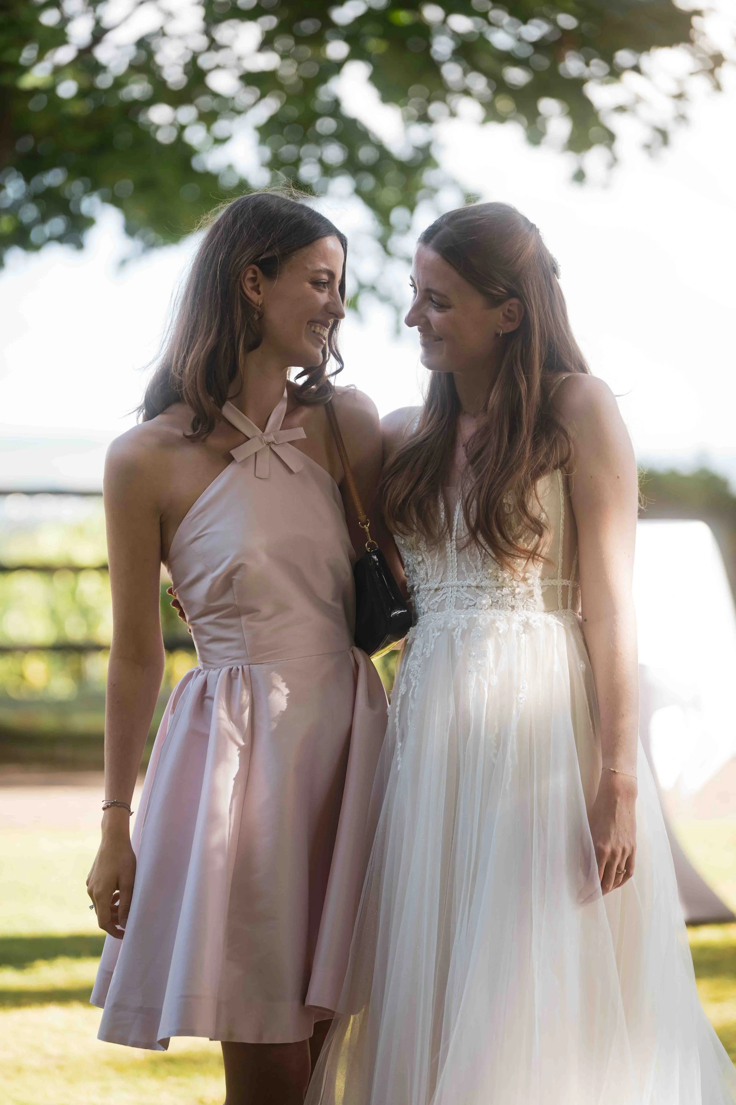 Zwei Frauen bei einer Hochzeit, die sich ansehen und lächeln, im Freien mit Bäumen im Hintergrund.
