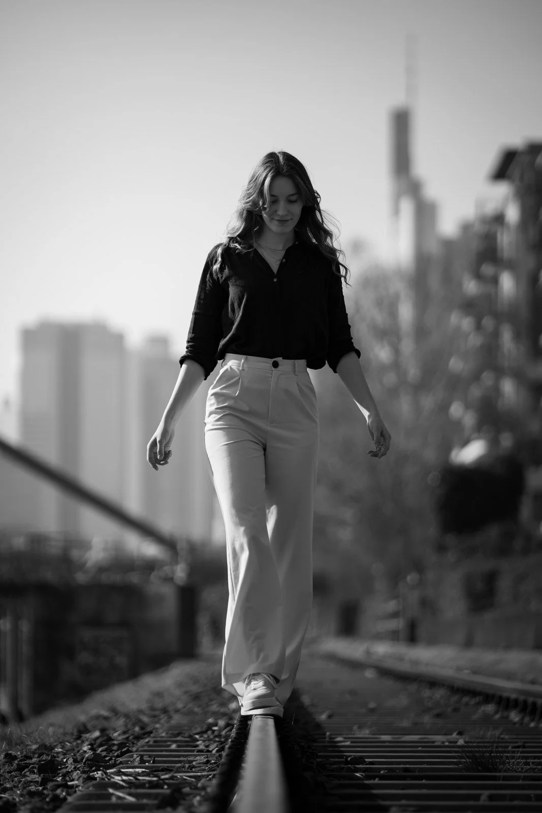 Eine Frau geht auf einem Eisenbahnstreifen in einer urbanen Umgebung, schwarz-weiße Aufnahme.