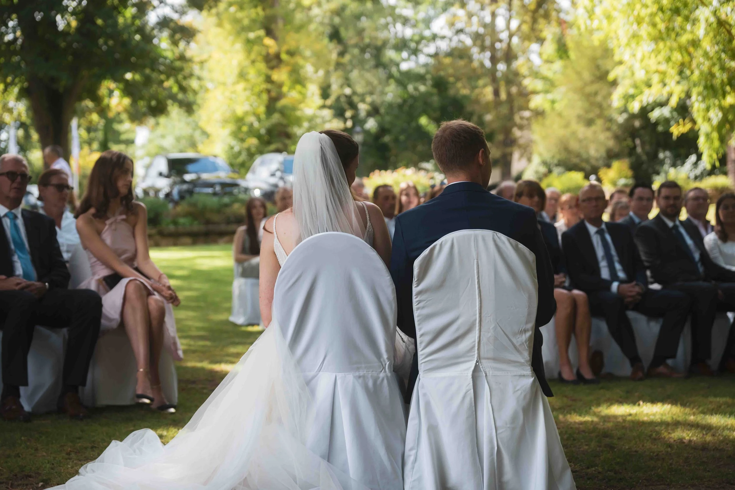 Ein Brautpaar sitzt auf weißen Stühlen bei einer Hochzeitszeremonie im Freien, umgeben von Gästen, im Hintergrund Bäume und geparkte Autos, sonniges Wetter.