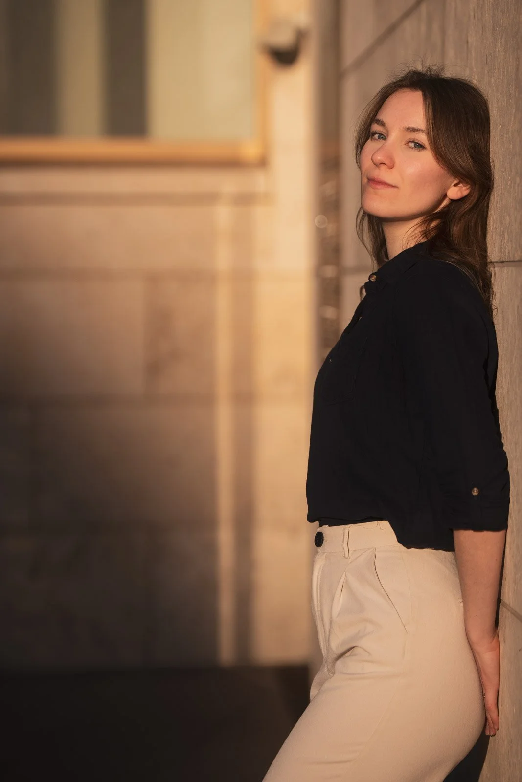 Junge Frau lehnt an einer Wand in einem modernen Innenraum, trägt beige Hosen und eine schwarze Bluse, mit braunen Haaren, lächelnd.