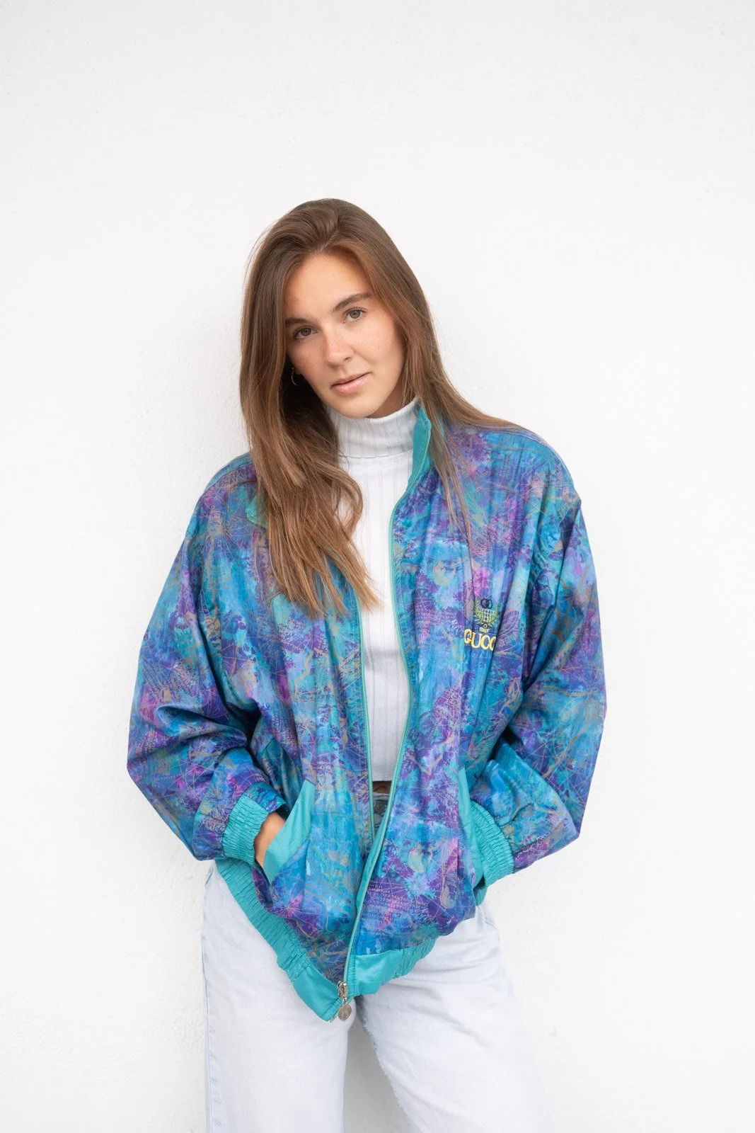 Junge Frau trägt eine bunte, sportliche Jacke und weiße Hose, steht vor weißer Wand und schaut in die Kamera.