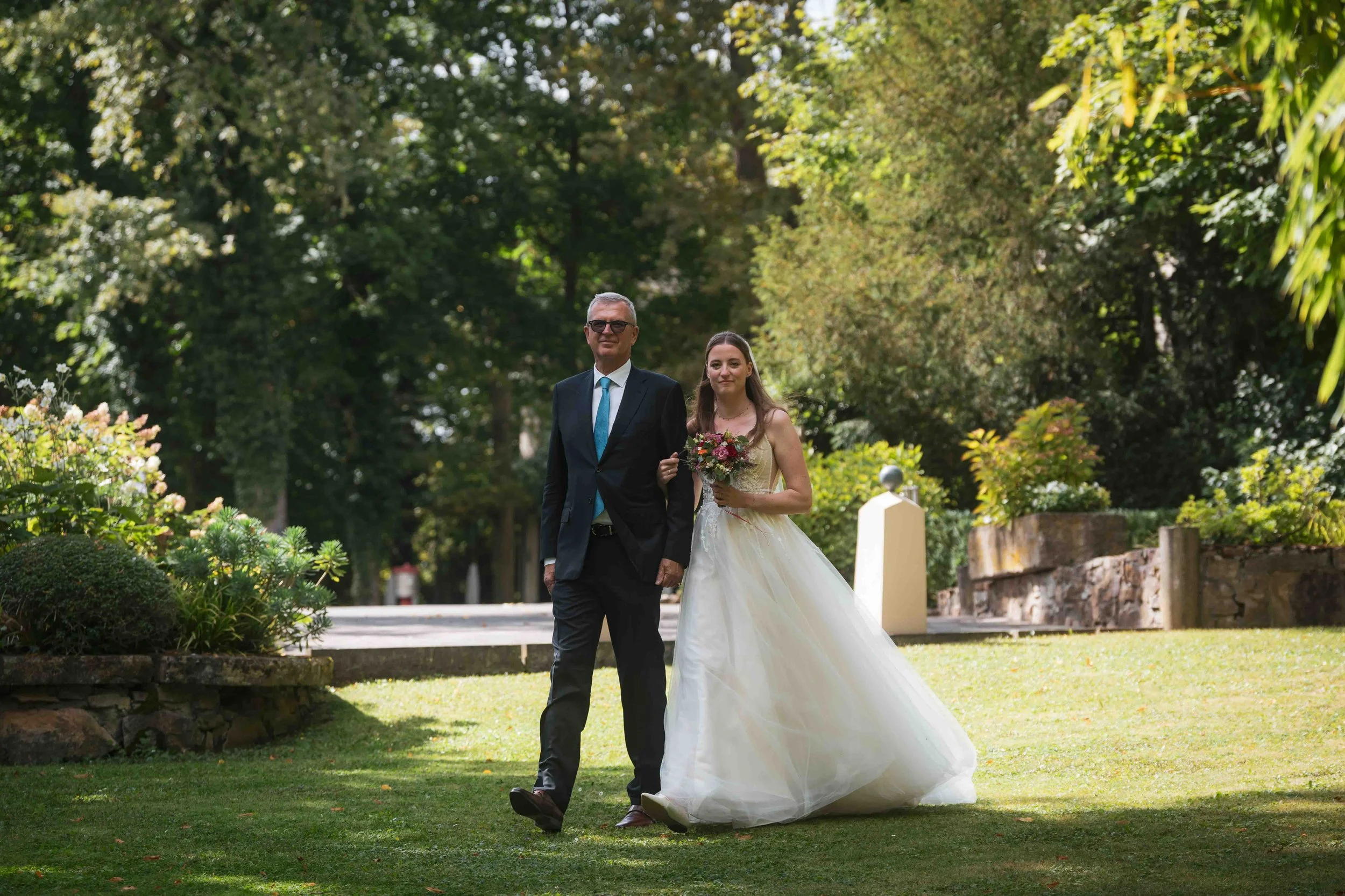 Braut im weißen Kleid wird von Mann in dunklem Anzug im Garten begleitet, sie hält einen Blumenstrauß.