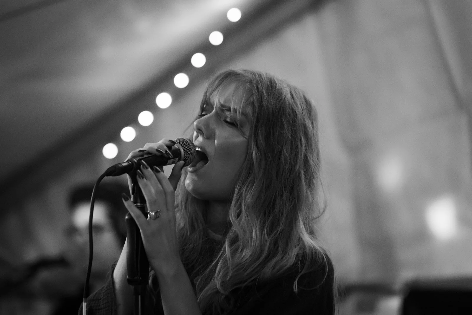 Schwarz-Weiß-Foto einer singenden Frau mit langen, lockigen Haaren bei einer Performance, im Hintergrund Lichter.