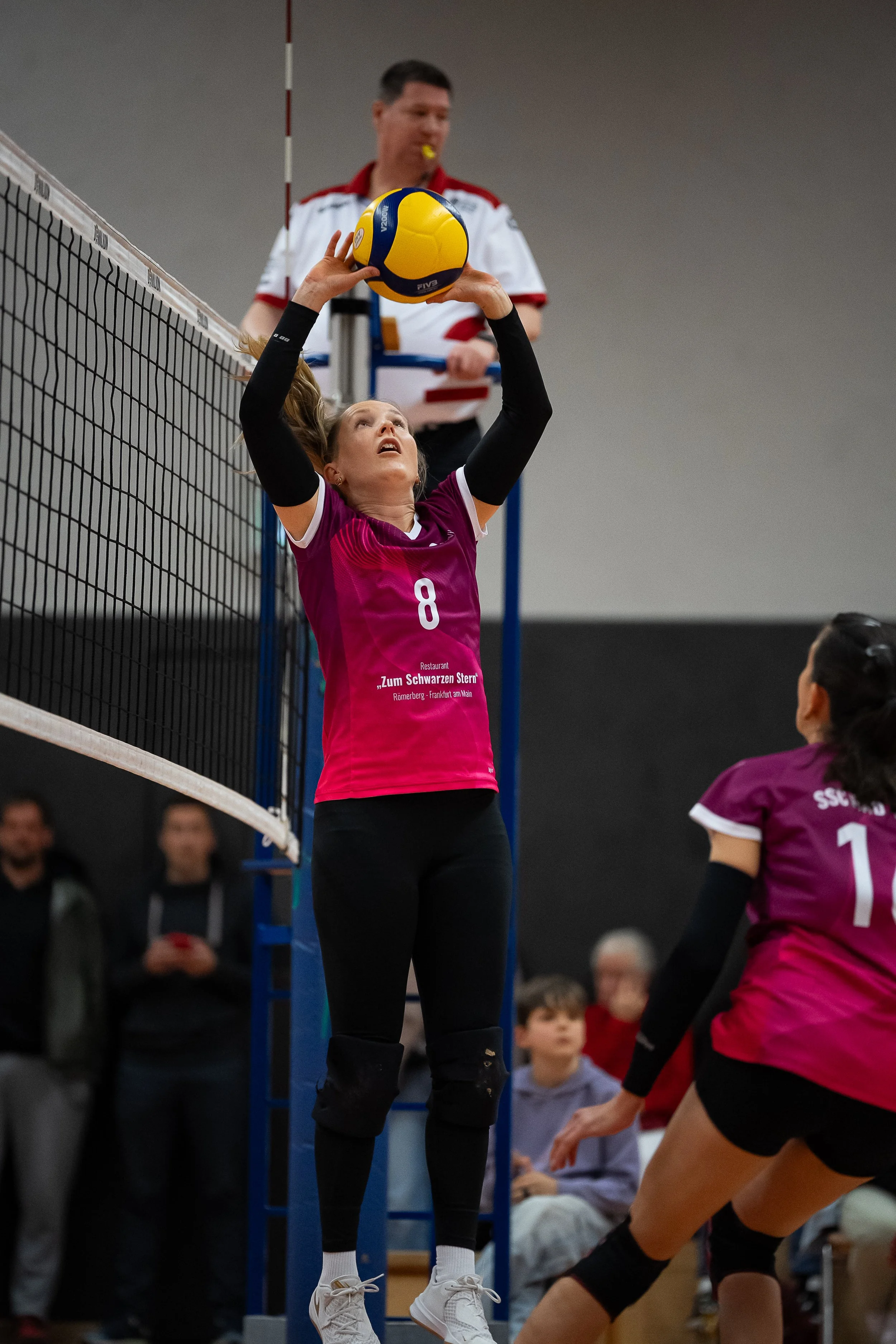 Eine Volleyballspielerin in Pink hebt den Ball über dem Netz, während eine Mitspielerin auf der rechten Seite vorbereitet ist, den Ball zu treffen. Im Hintergrund beobachtet ein Schiedsrichter mit gelbem Pfannen den Spielverlauf, und einige Zuschauer