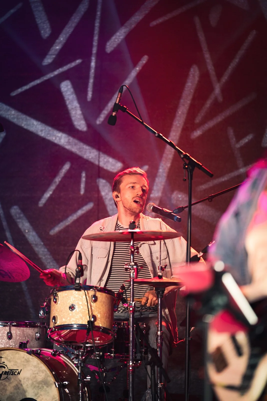 Ein junger Mann spielt Schlagzeug auf einer Bühne mit rotem und lila Licht, im Hintergrund ist eine abstrakte Wand mit Lichtstrahlen zu sehen.