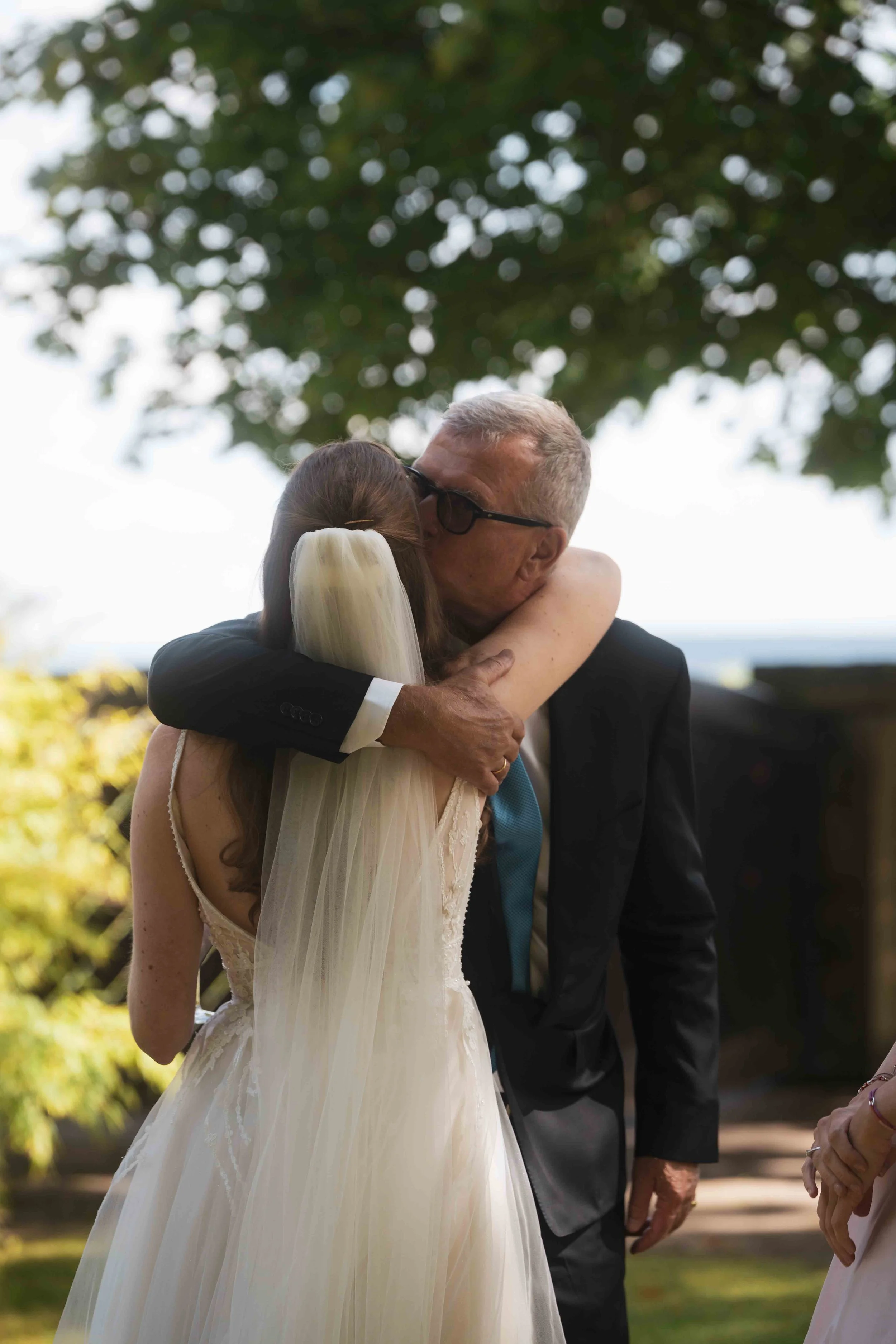 Ein älterer Mann in Anzug und eine junge Frau mit Hochzeitskleid umarmen sich. Der Mann trägt dunkle Brille und hat graue Haare. Das Bild ist im Freien mit Bäumen im Hintergrund aufgenommen.