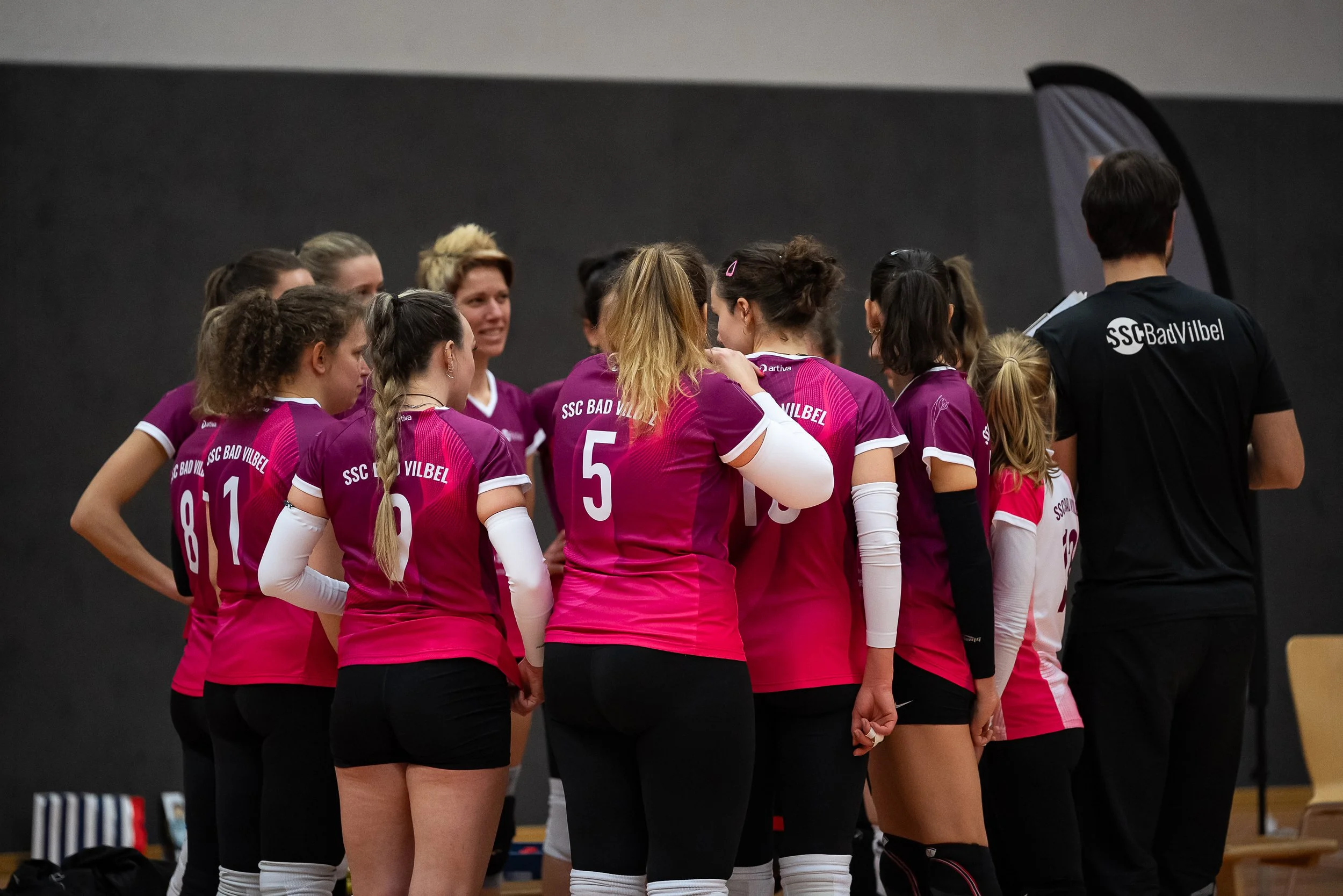 Eine Gruppe von Volleyballspielerinnen in pinken Trikots und weißen Armbändern während eines Teams er Keywordmerksamkeit, die sich in einem schwarzen Raum versammelt, möglicherweise vor einem Spiel oder einer Pause.