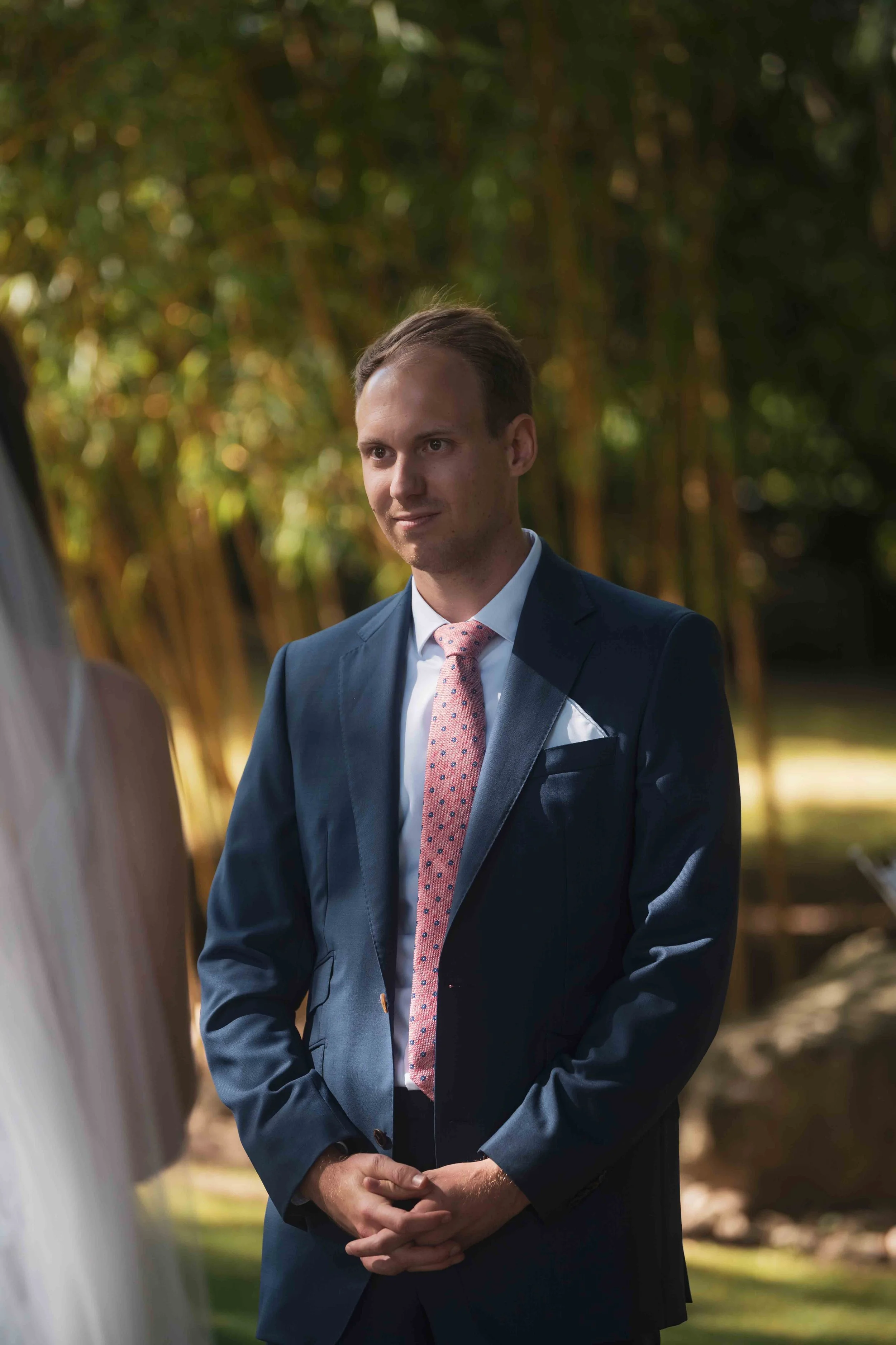 Ein Mann in formeller Kleidung beim Outdoor-Ereignis, vermutlich bei einer Hochzeitszeremonie, im Hintergrund grüne Bäume und Sonnenlicht.