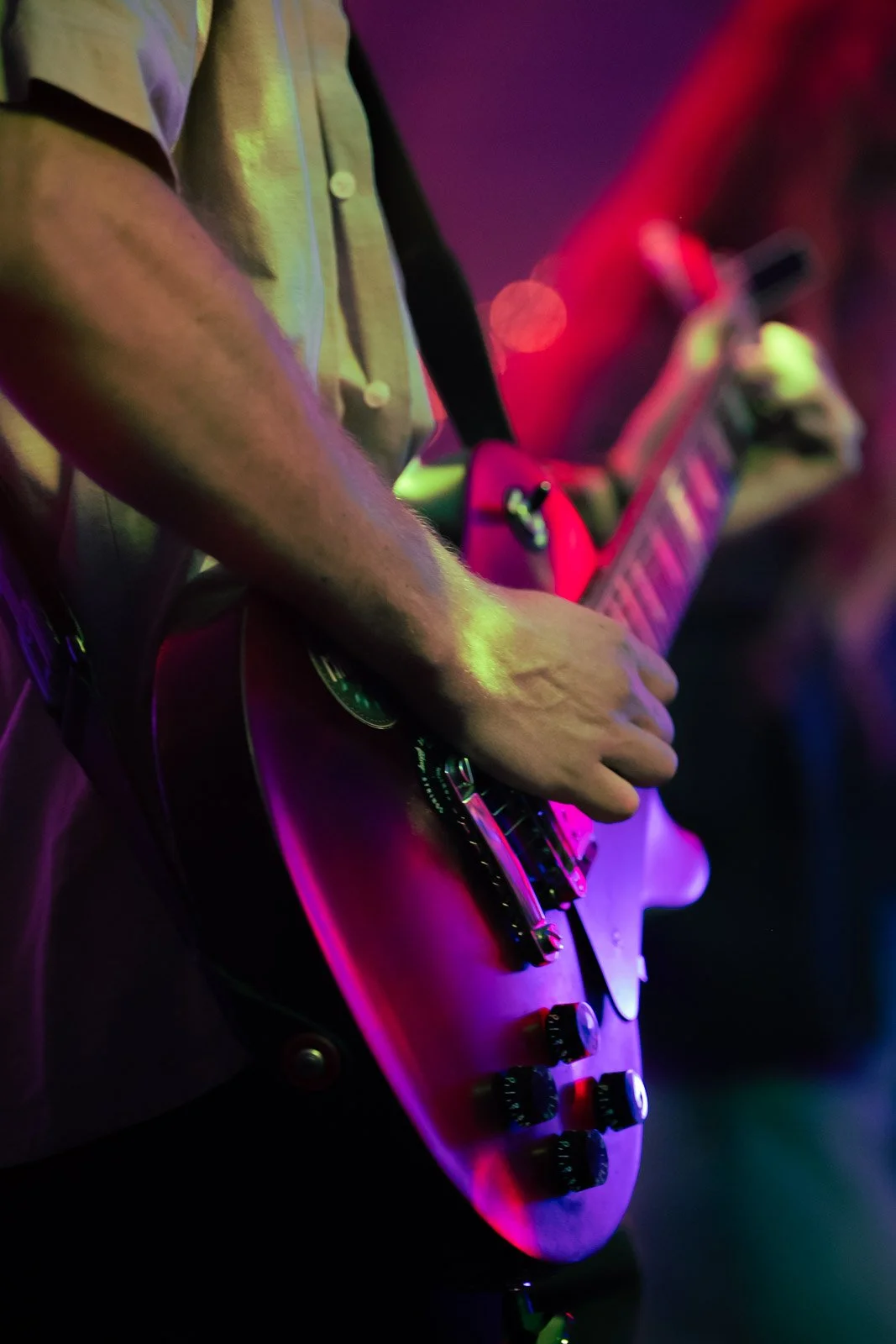 Nahaufnahme einer Person, die einen E-Gitarrenkopf in einer dunklen, farblich beleuchteten Umgebung spielt.