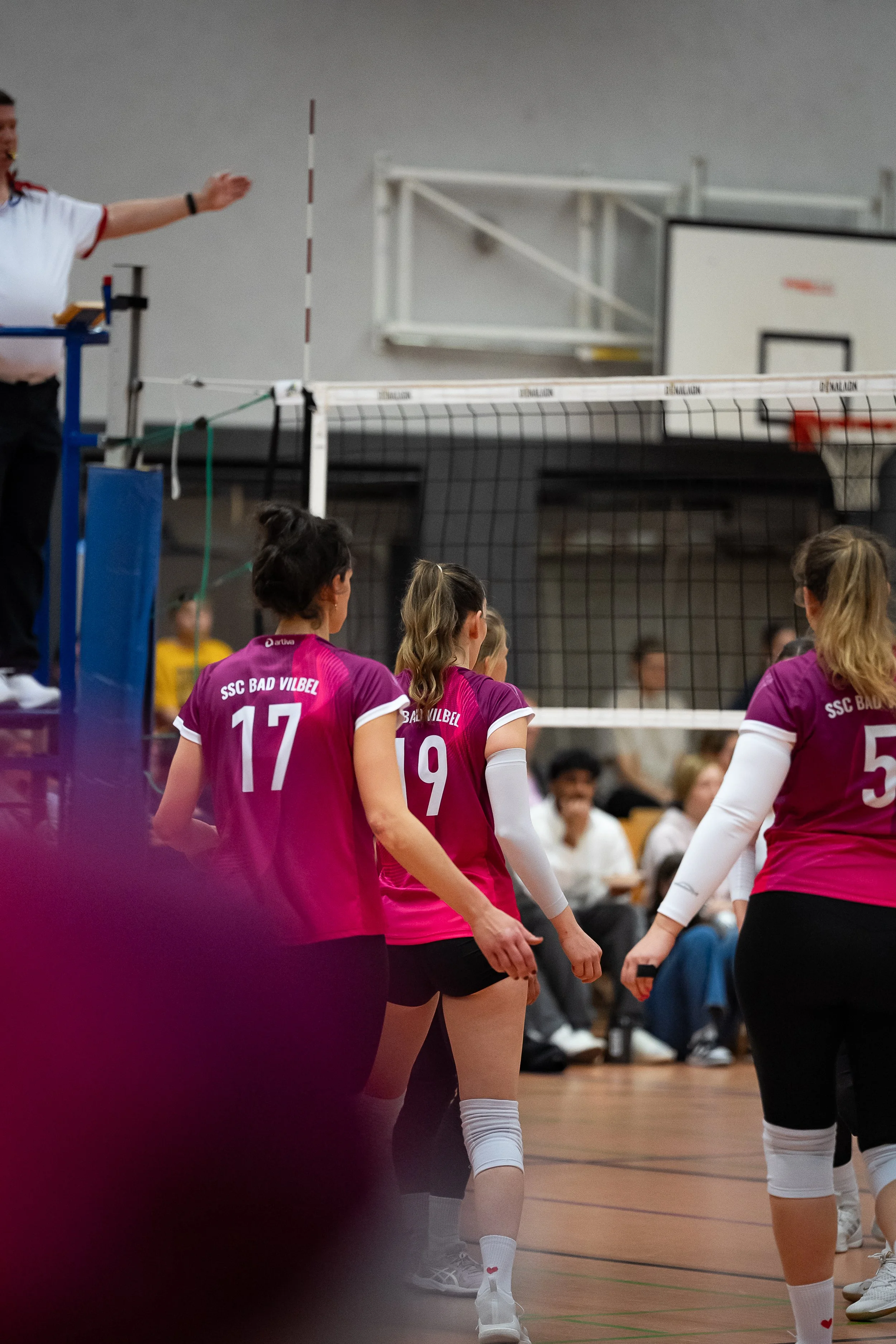 Volleyballspielerinnen in pinken Trikots stehen auf dem Spielfeld während eines Spiels im Innenraum, mit Zuschauern im Hintergrund.