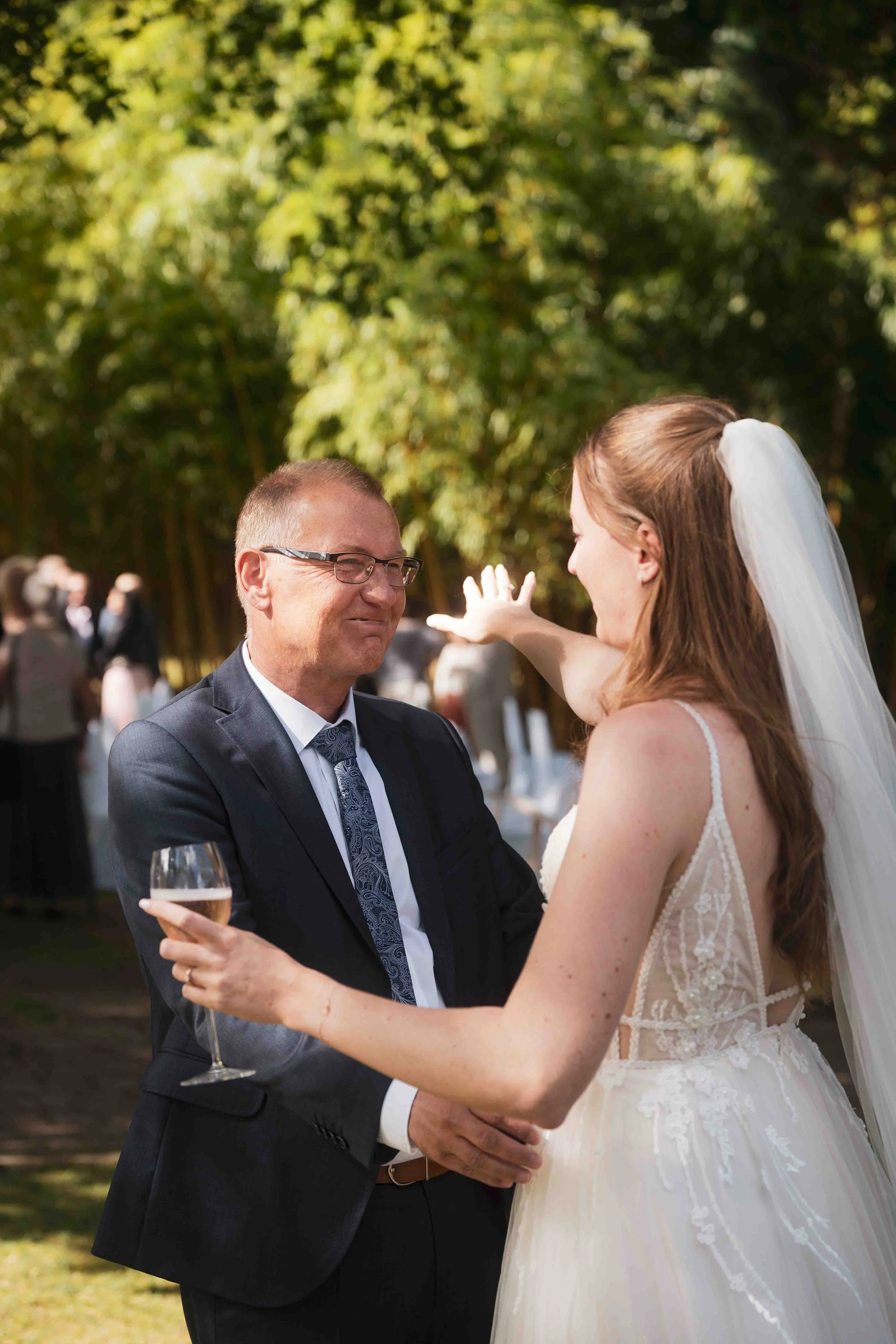 Hochzeit im Freien: Ein Mann in Anzug und eine Braut in Weiß feiern gemeinsam bei Sonnenlicht, umgeben von Bäumen.