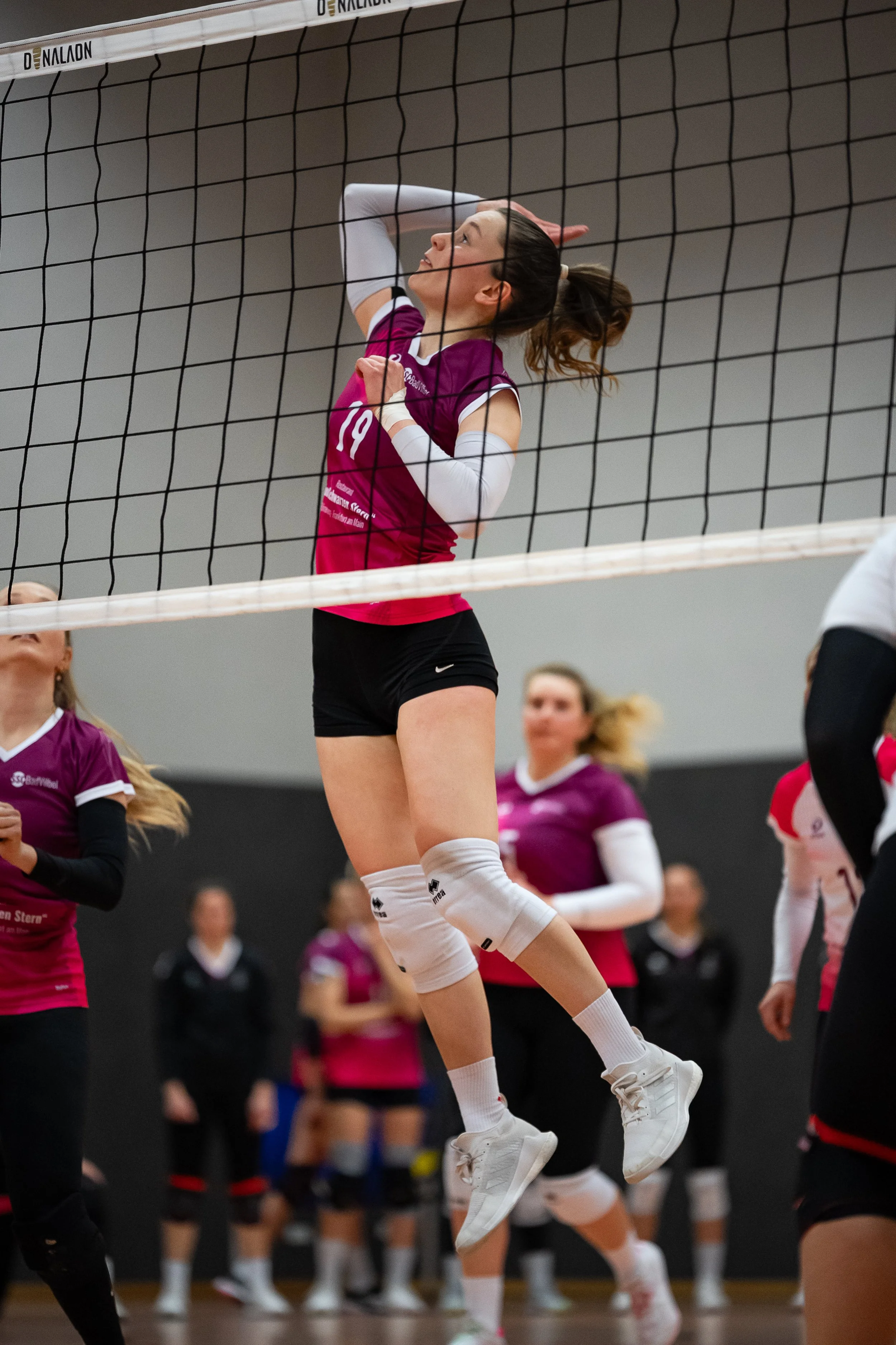 Volleyballspielerin springt am Netz während eines Spiels, andere Spielerinnen im Hintergrund.