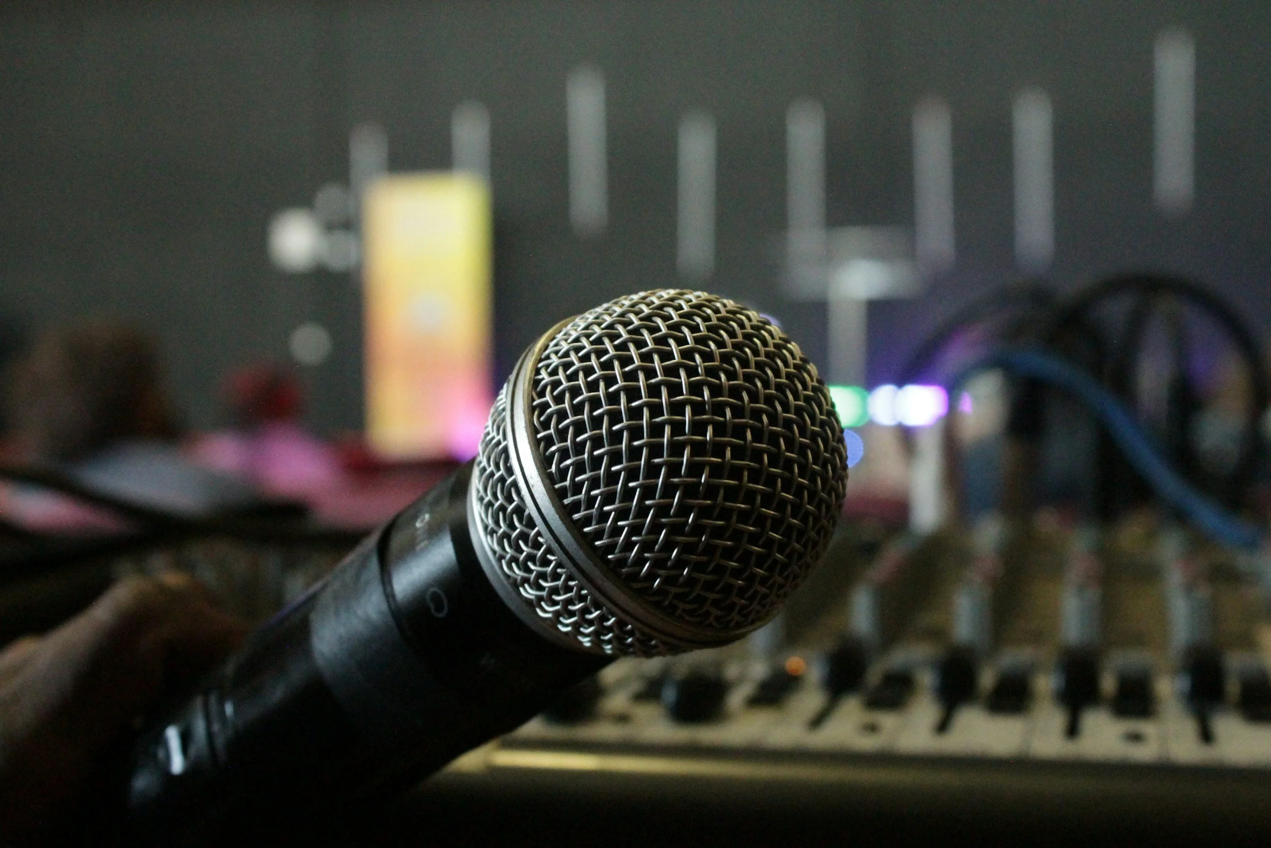 Close-up of a microphone in front of audio mixing equipment with blurred colorful signs in the background.