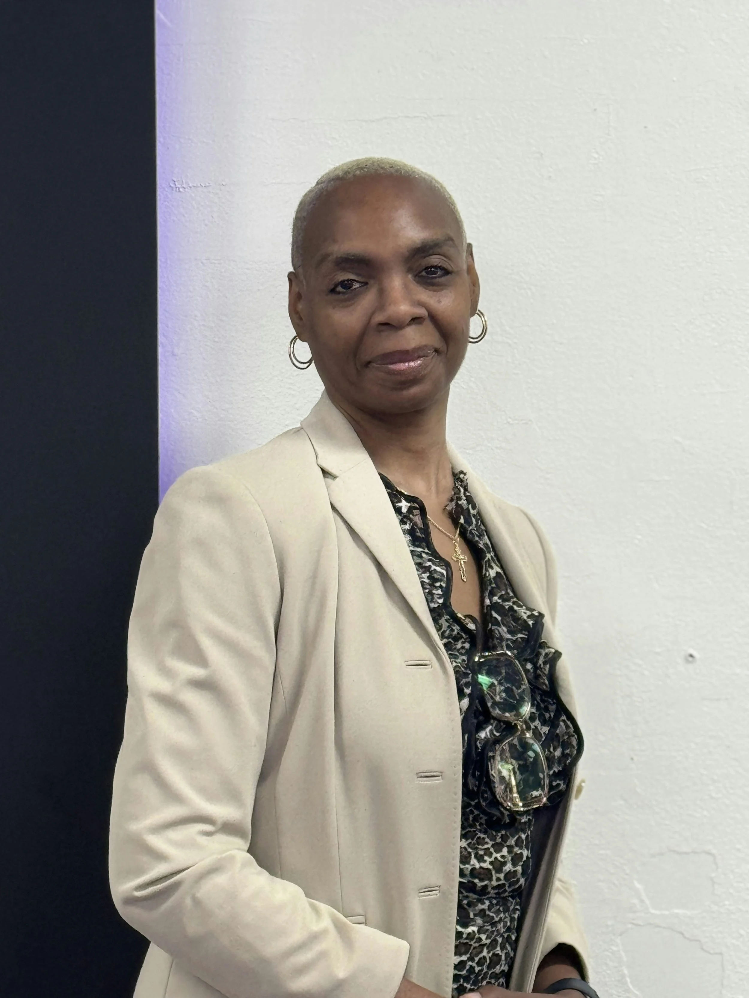 A woman with short, gray hair wearing a beige blazer over a leopard print blouse, accessorized with glasses hanging from her shirt, gold earrings, and a necklace with a cross pendant, standing against a plain wall.