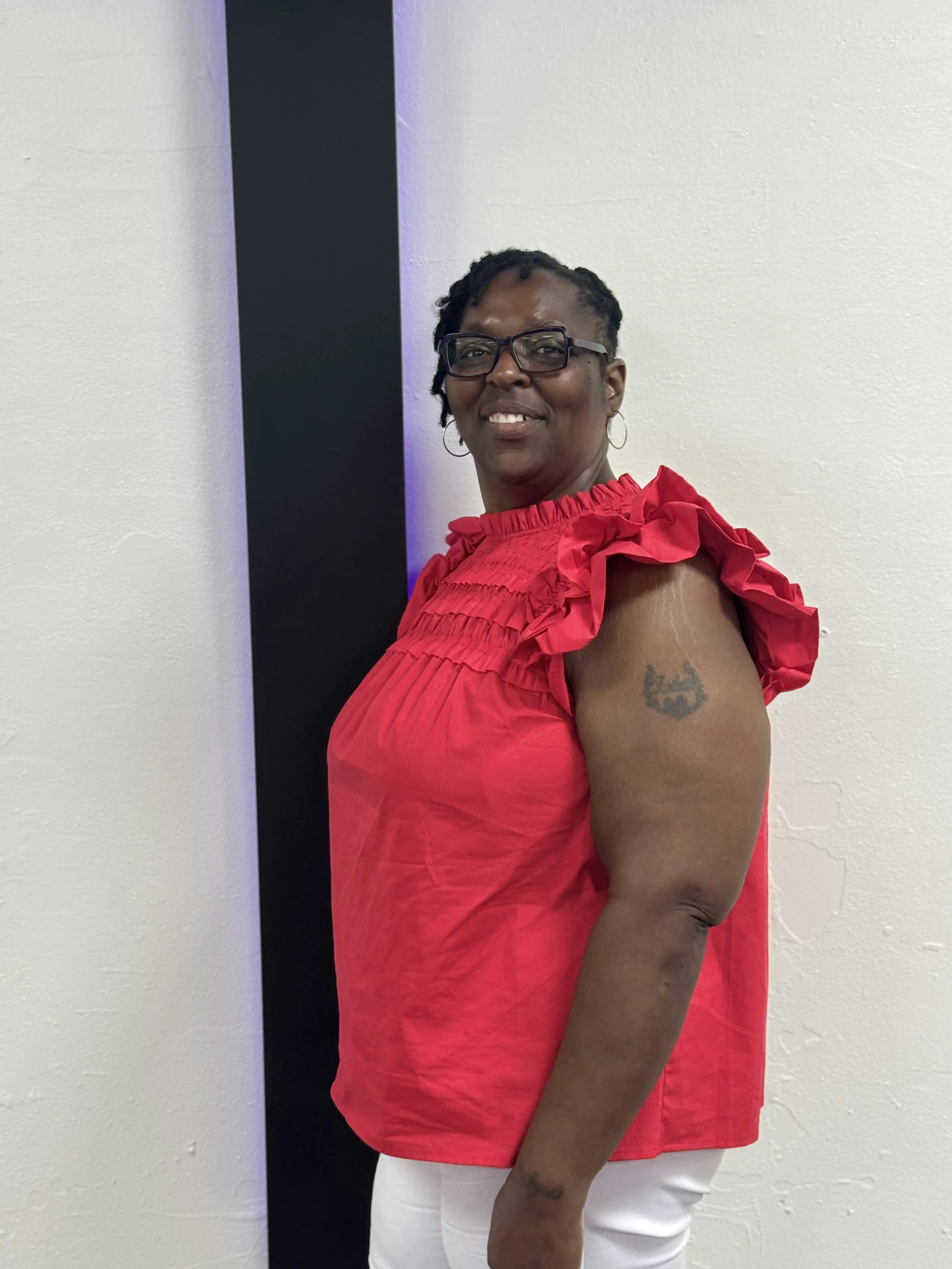 A woman smiling, wearing glasses and a red ruffled sleeveless top, stands against a white wall with a black vertical stripe.