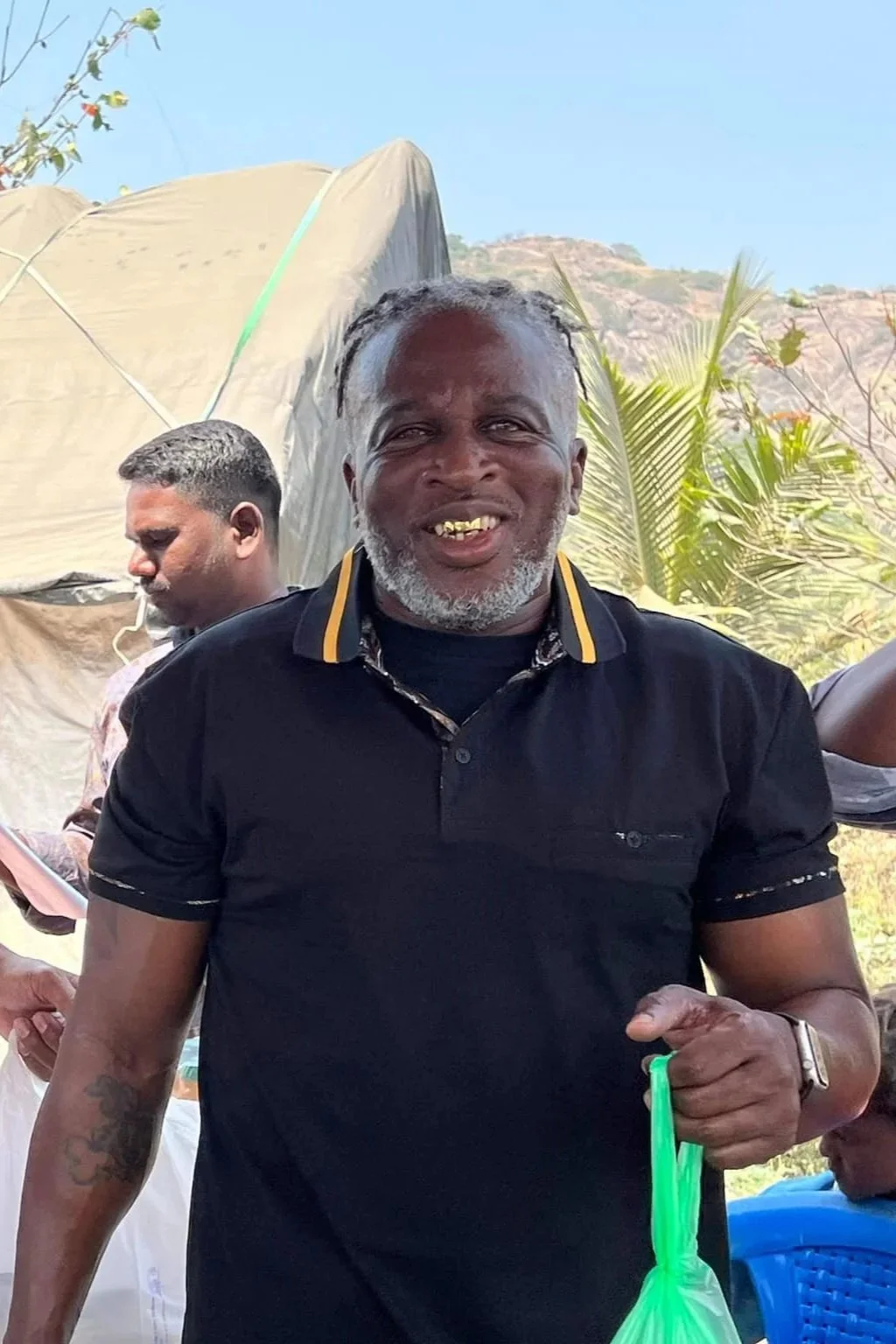 A smiling man with gray hair and a beard, wearing a black polo shirt, holding a green plastic bag, outdoors with mountains and plants in the background.