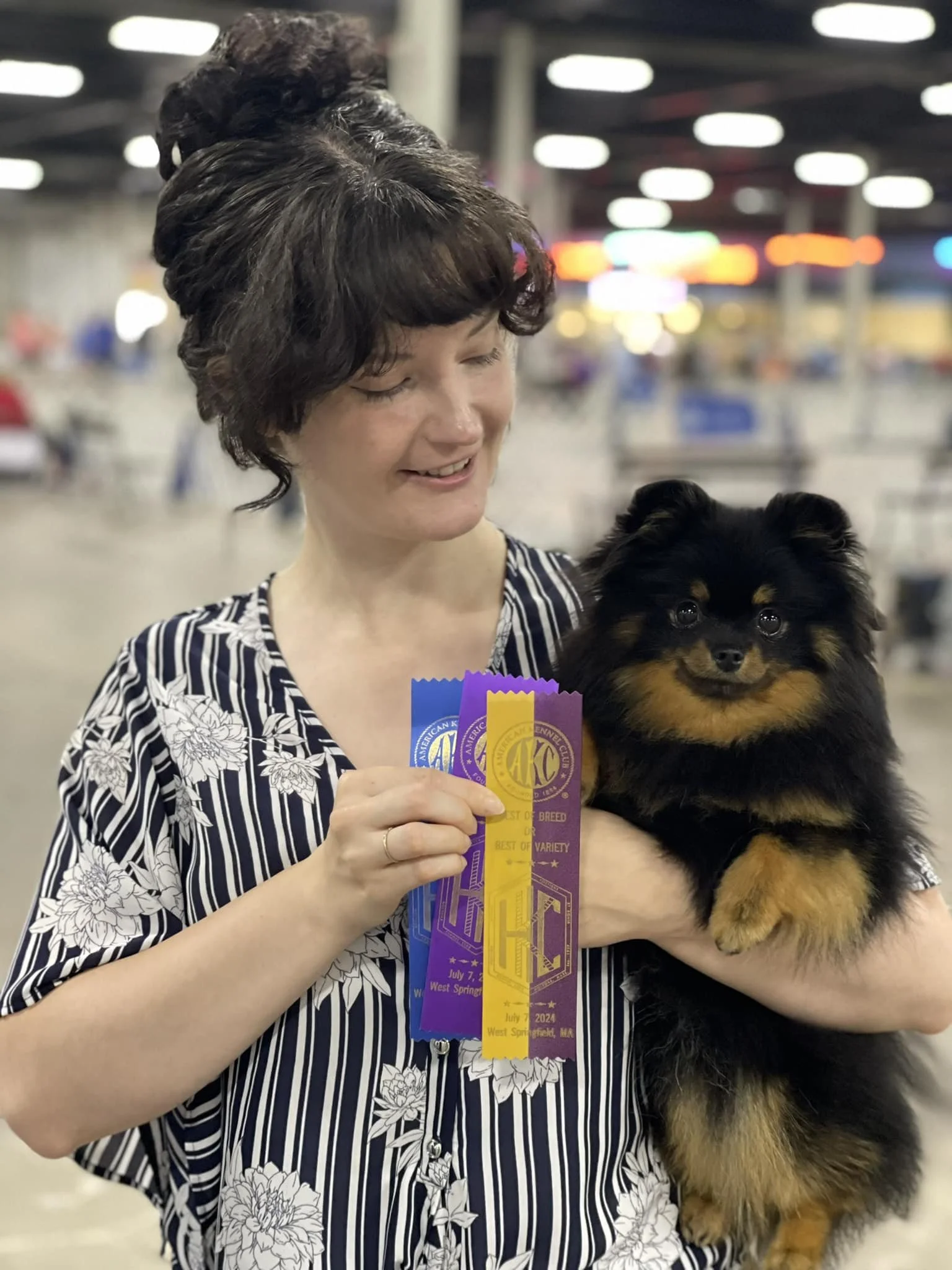 A woman with a blue and white striped blouse stands at a dog show holding her black and tan Pomeranian in her left arm, while holding up multi colored ribbons in her right hand, looking lovingly at her dog.