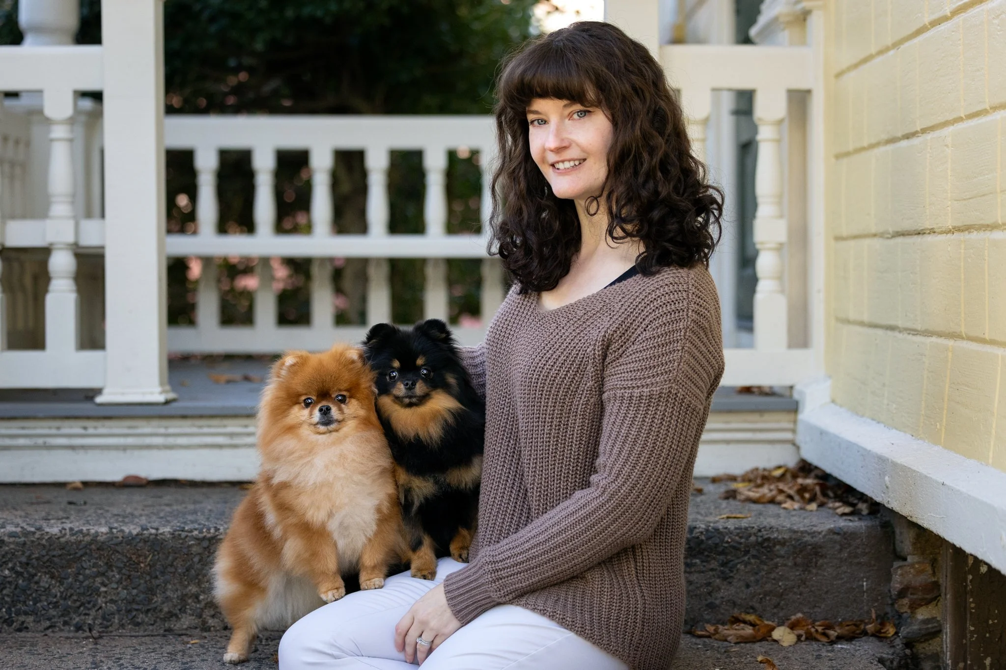 A woman with pale skin, brown curly hair and blue eyes wearing a brown knit sweater and white pants sits on a stone colonial porch with her two Pomeranians with their front two legs on the person's thigh, everyone looking at the camera.