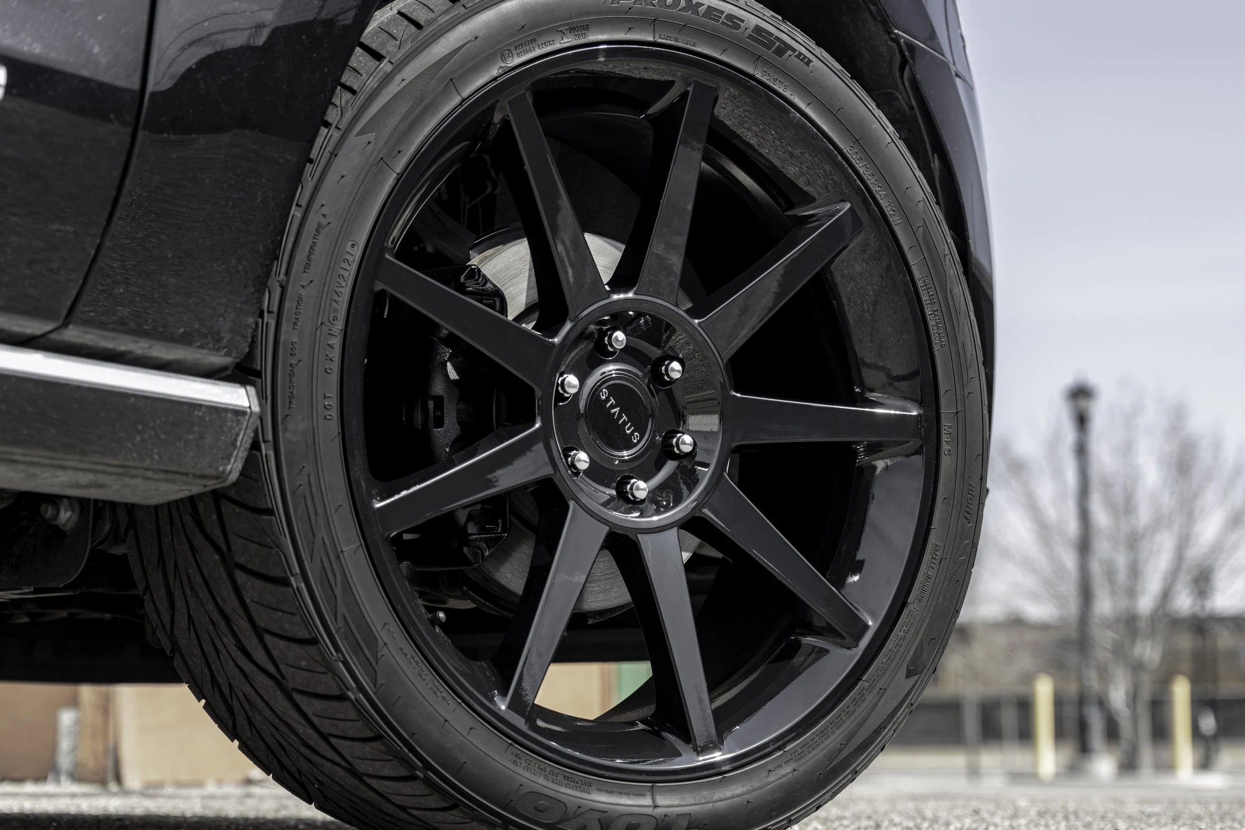 Close-up of a black alloy wheel with a tire, part of a black vehicle, outdoors in a parking lot with trees and a streetlamp in the background.