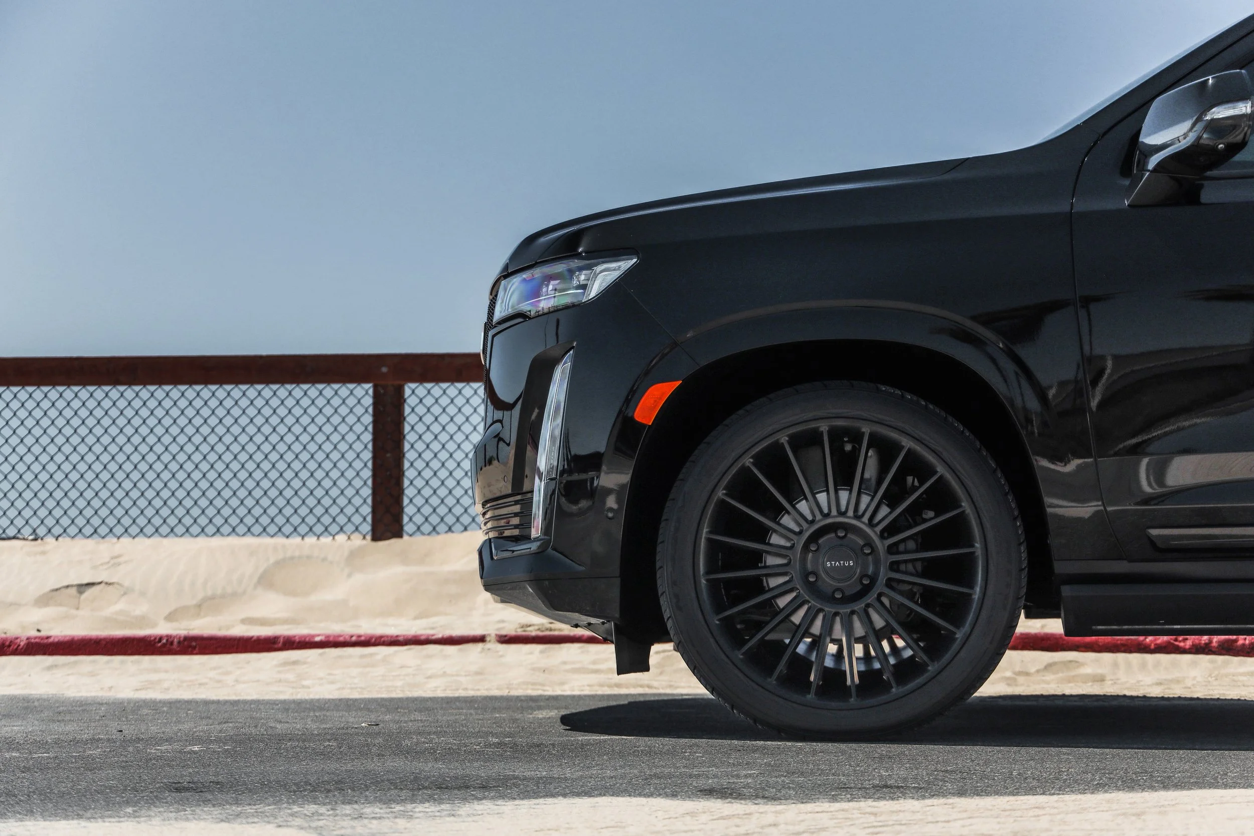 Front of a black SUV parked on a concrete surface near a sand area with a metal fence in the background.