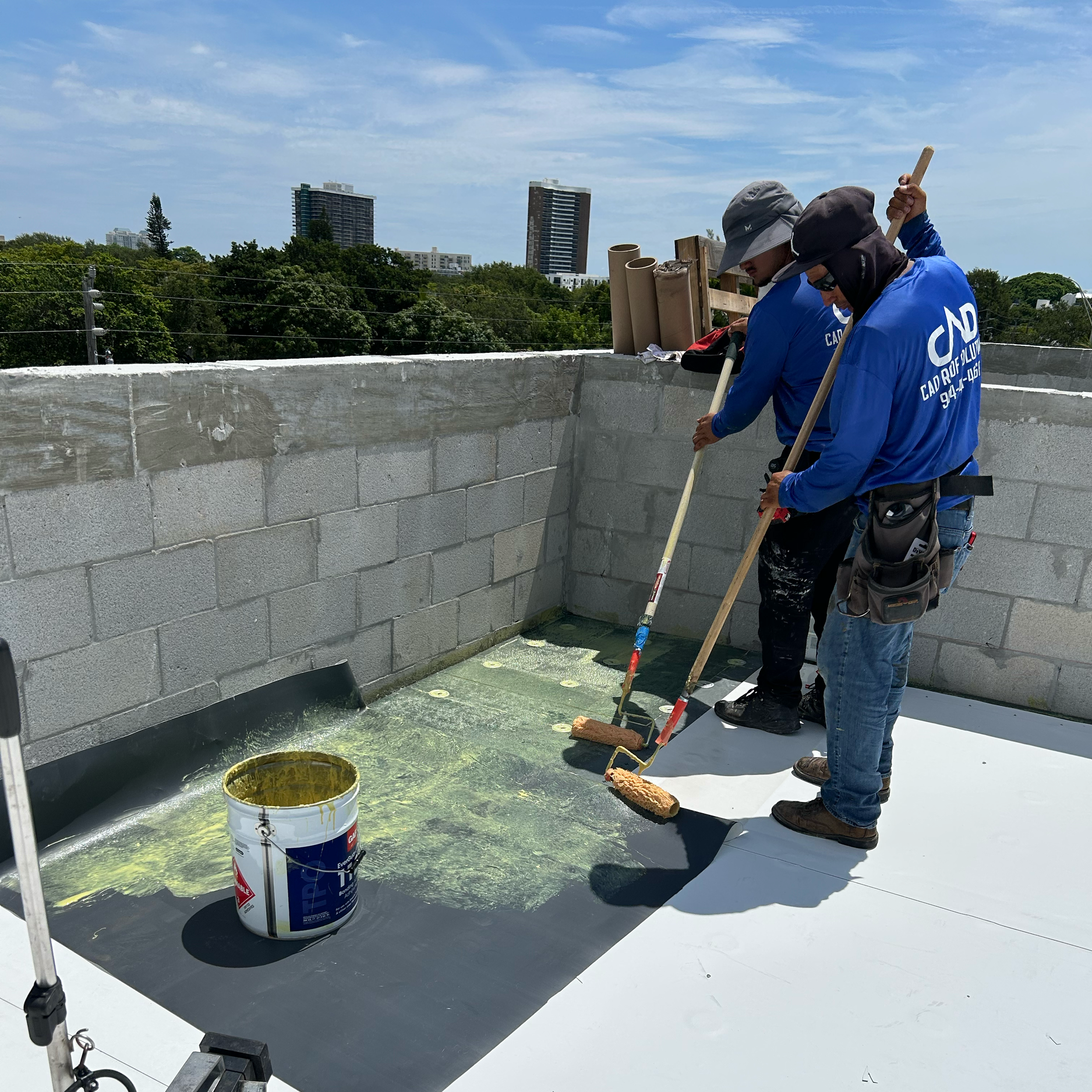 Two workers applying waterproofing membrane on a rooftop with paint rollers, with a bucket of waterproofing material nearby, in an urban setting with high-rise buildings in the background.