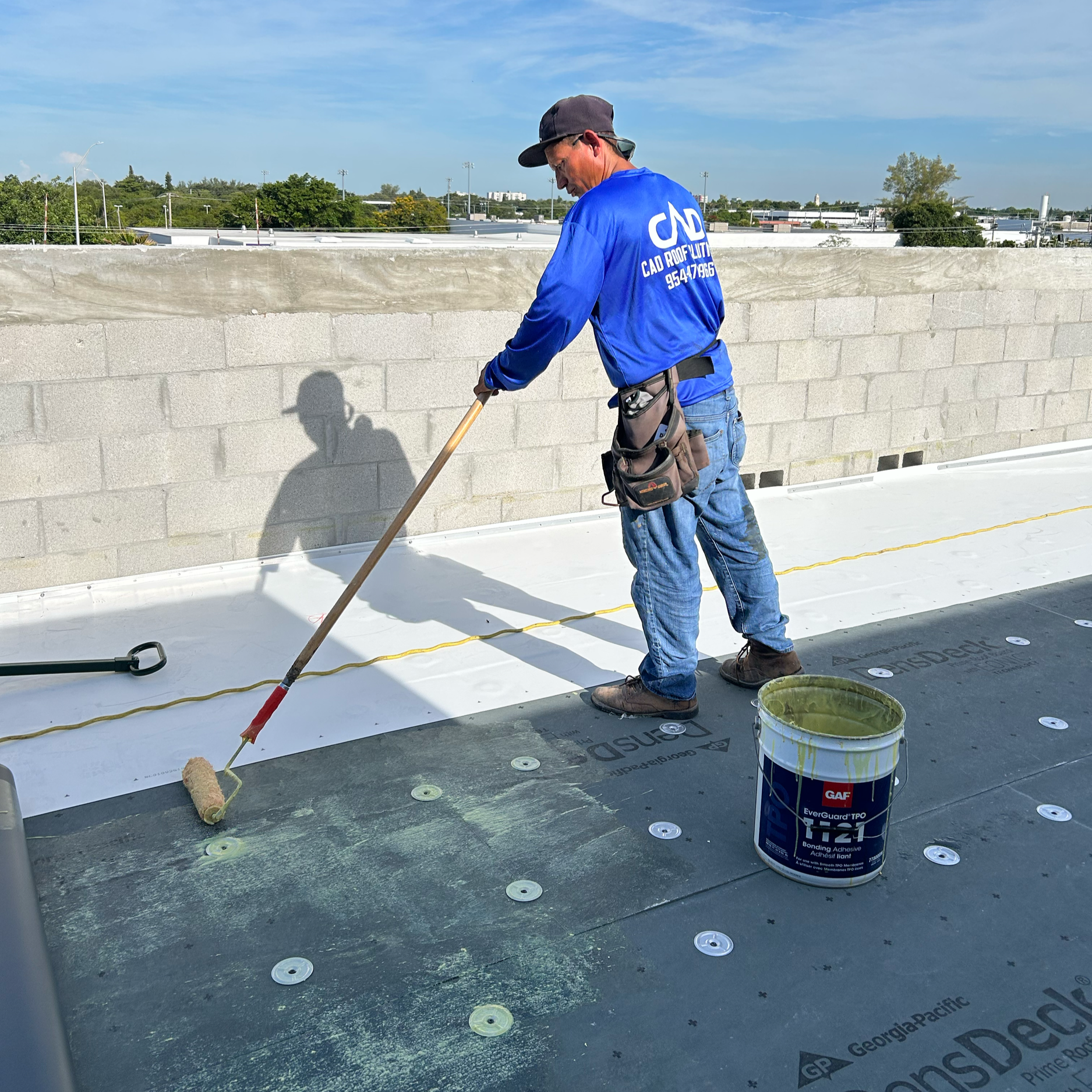 A worker on a roof using a roller brush to apply adhesive to a roofing membrane. He is wearing a blue shirt, jeans, a cap, and has a tool belt. There is a bucket of adhesive nearby.