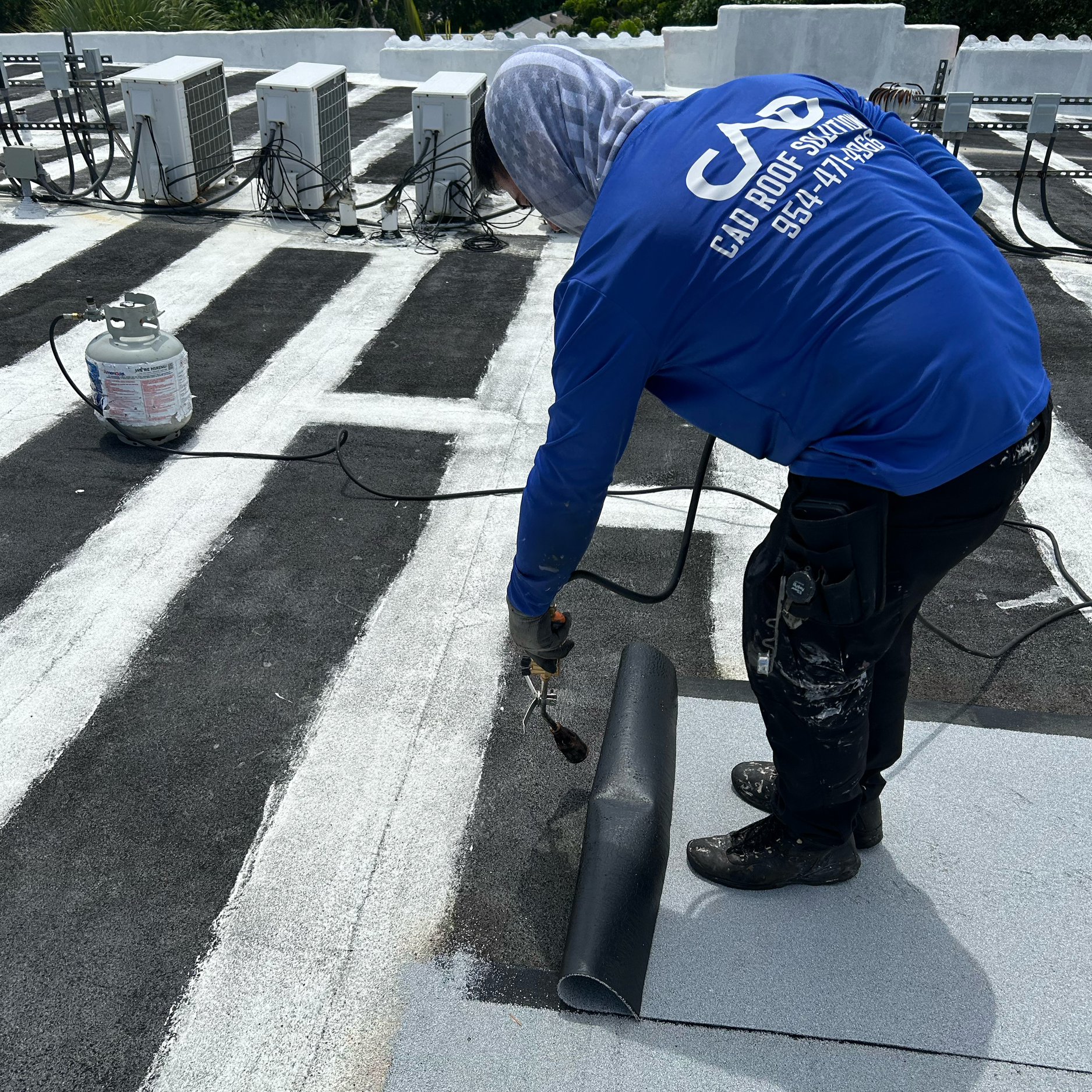 A worker in a blue jacket and gloves coating a rooftop with a white reflective coating using a spray tool, with additional coating supplies and air conditioning units in the background.
