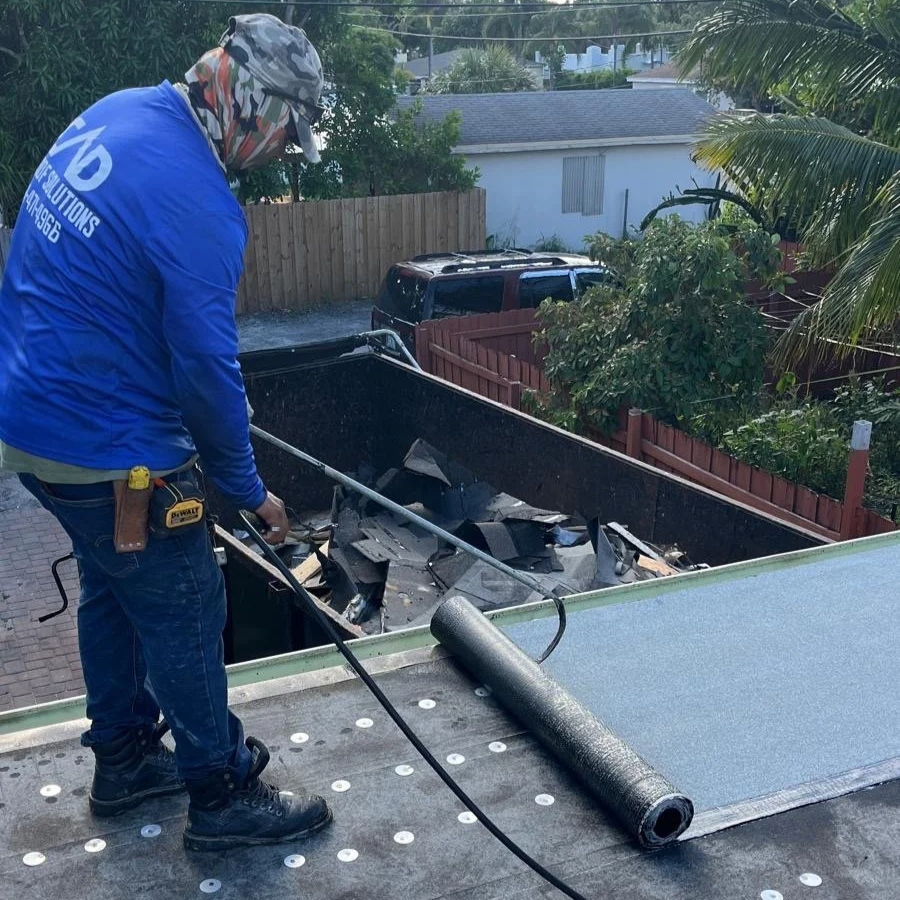 A worker installing or repairing a flat roofing membrane on a building's roof, with a roll of roofing material nearby.