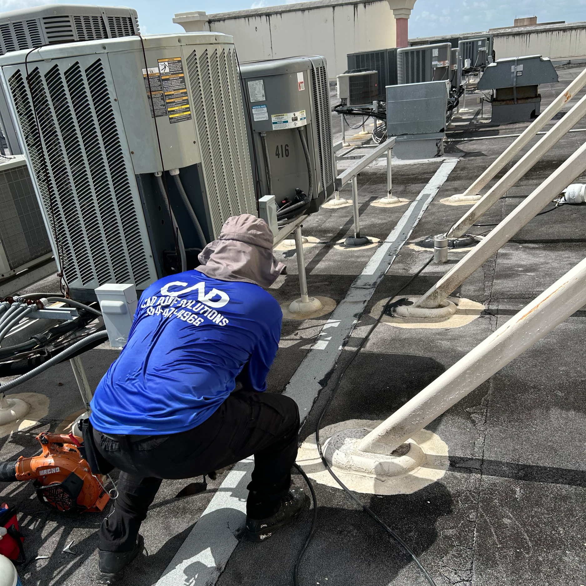 A technician kneels on a rooftop working with HVAC or electrical equipment, wearing a blue shirt and wide-brimmed hat, beside various rooftop units and support beams.