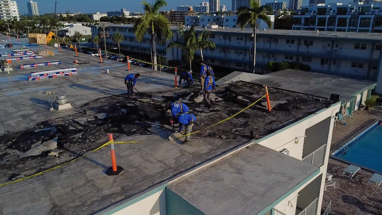 Workers repairing a burnt rooftop at a hotel in a sunny city, with palm trees and pool area nearby.