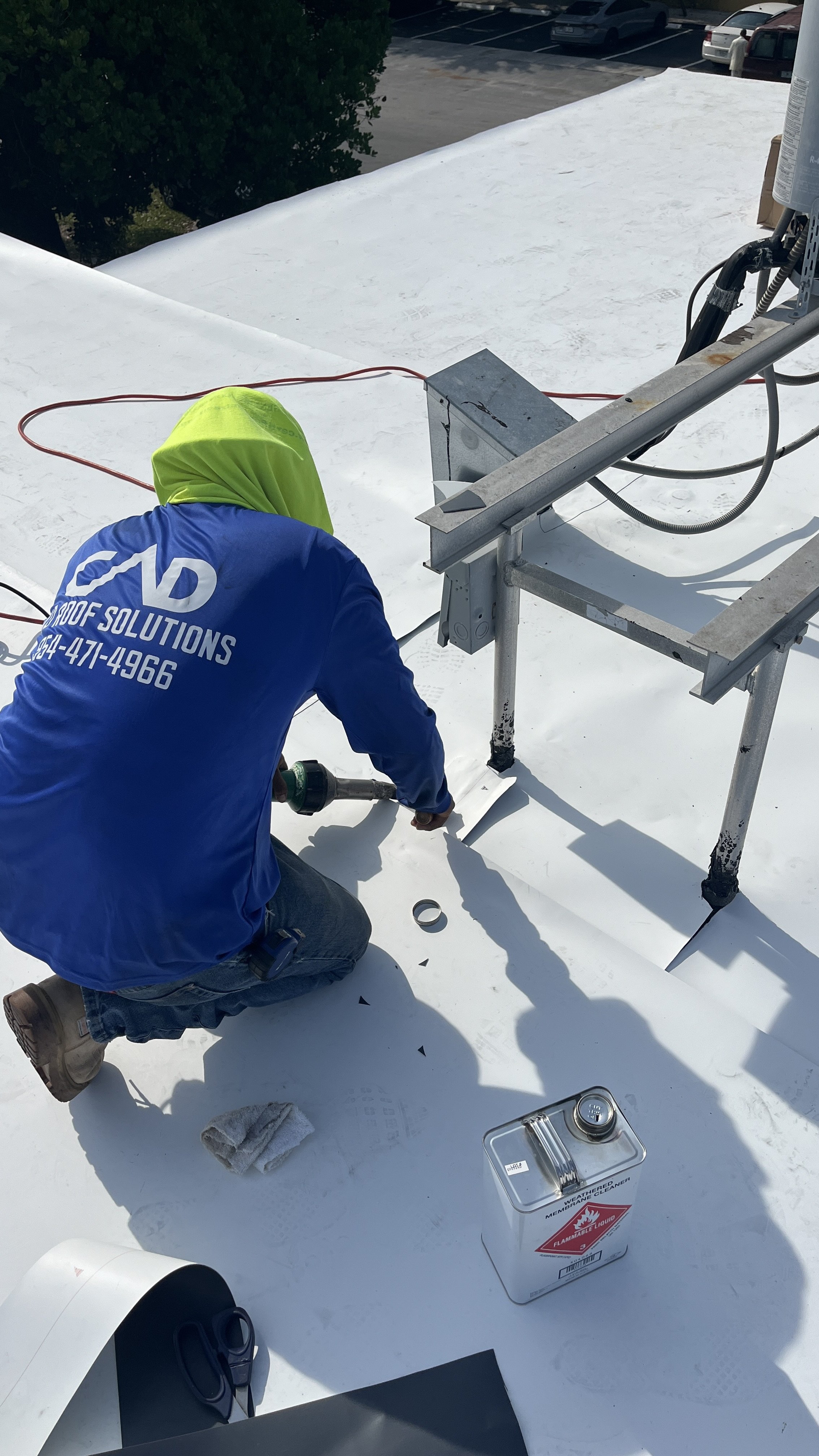 A worker with a yellow hood and blue shirt kneeled on a white rooftop, installing or repairing a solar panel or reflective roofing material. The worker is pneumatic or electric tool, with a cloth and a can of weatherproof fluid nearby. A parking lot 
